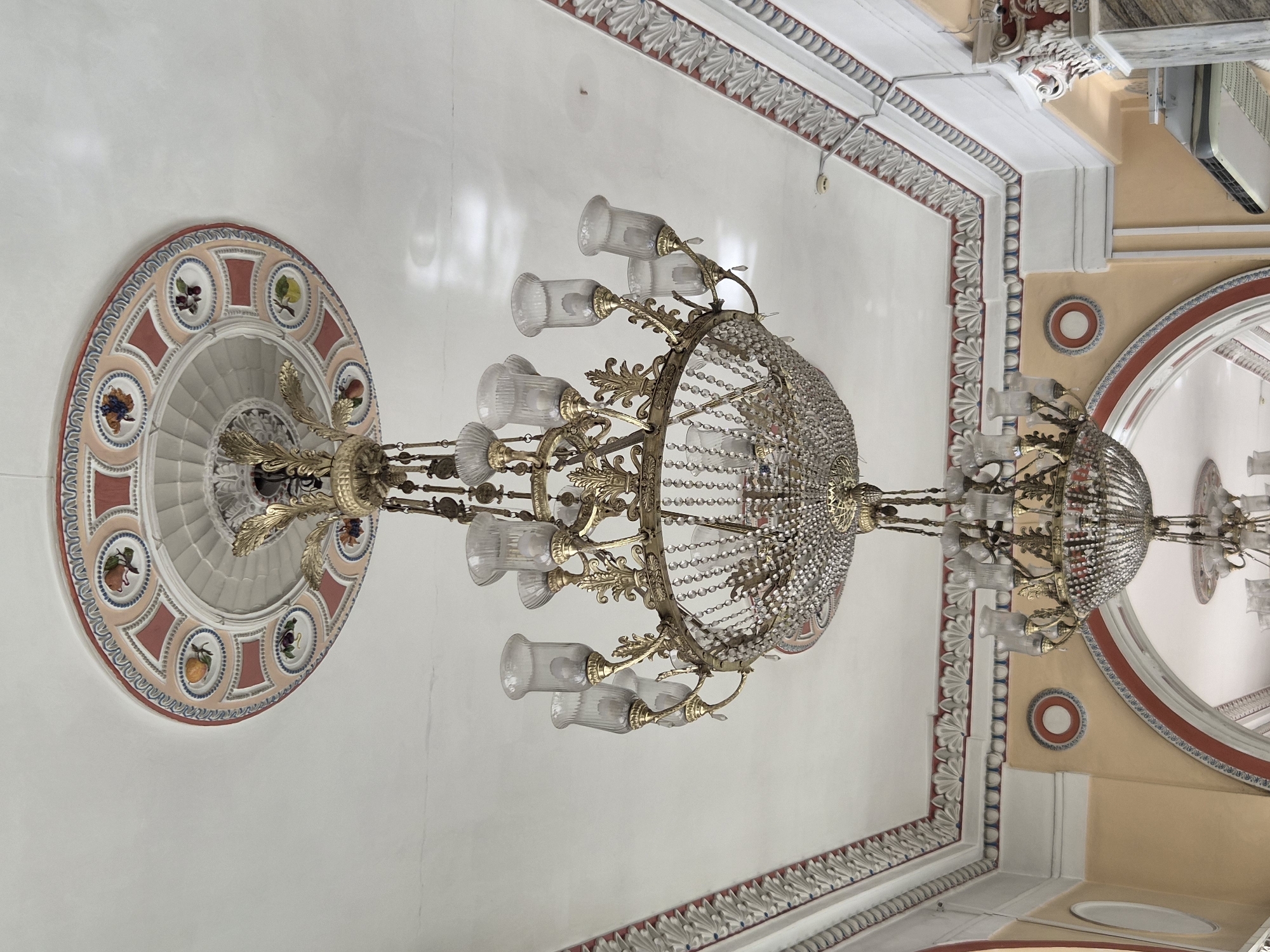 The image shows a view of the ceiling and upper walls of a room, featuring two ornate chandeliers. The ceiling is white with decorative molding that includes fruit motifs on a pink and blue background. The larger chandelier, made of gold-colored metal and crystal, hangs prominently in the center. Below it, a second, smaller chandelier is partially visible. The walls are painted a pale yellow with red and blue decorative accents and arched doorways. This scene is located in the Hall of Receptions of the Palace of the Republic in Minsk, Belarus.