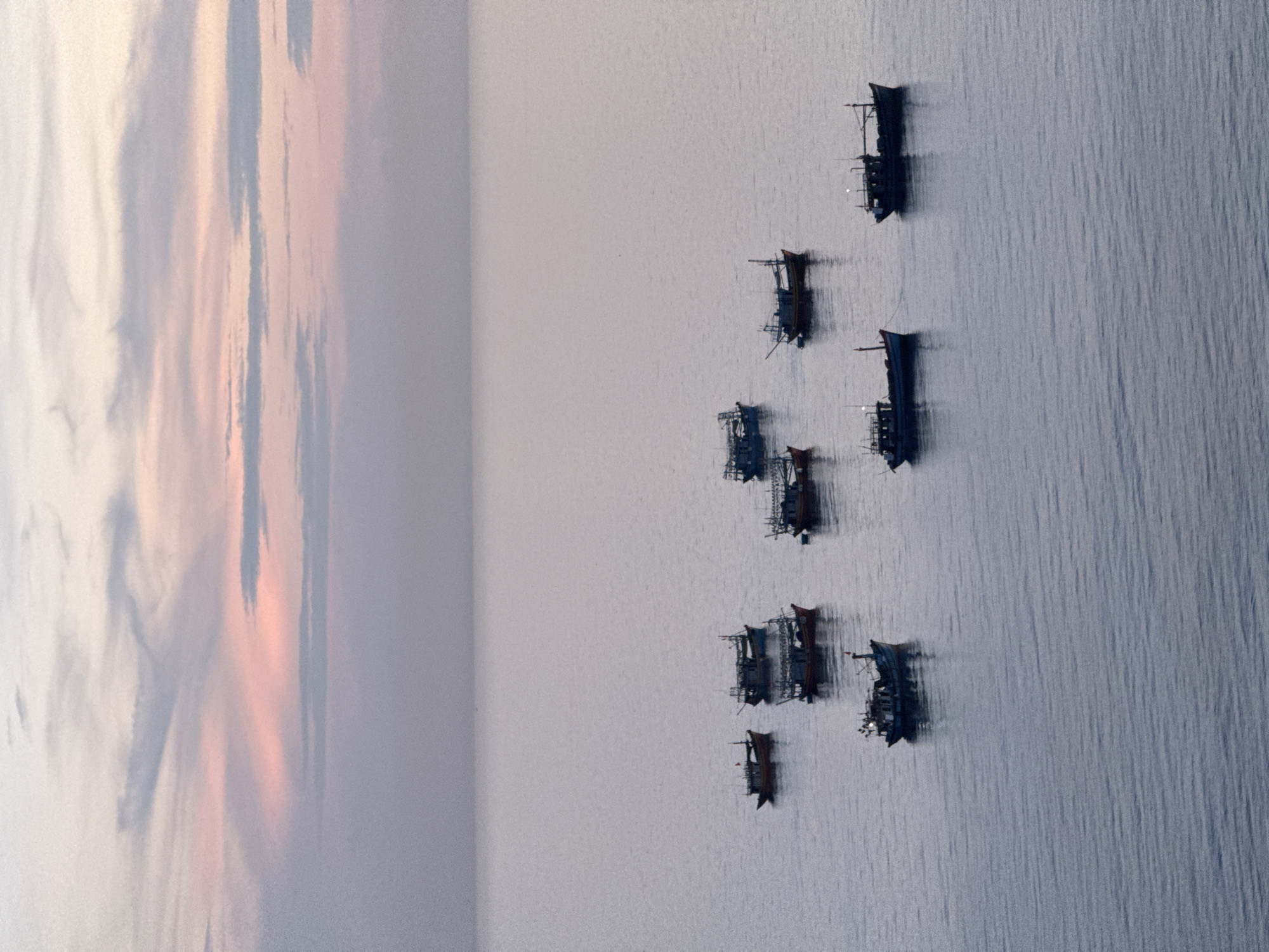 This is an aerial, high-angle, full shot of a calm, deep blue ocean with a pastel-colored sky at dusk or dawn. The sky transitions from pale pink and lavender hues at the horizon to a muted grey further up. Scattered across the water are approximately ten fishing boats of varying sizes, mostly dark in color, with some appearing to be anchored. The boats are positioned at different distances from the viewer, creating a sense of depth. The overall scene is serene and tranquil, suggesting a peaceful moment at sea. This image could depict the waters surrounding Lý Sơn Island in Vietnam, a location known for its fishing communities and beautiful coastal scenery.