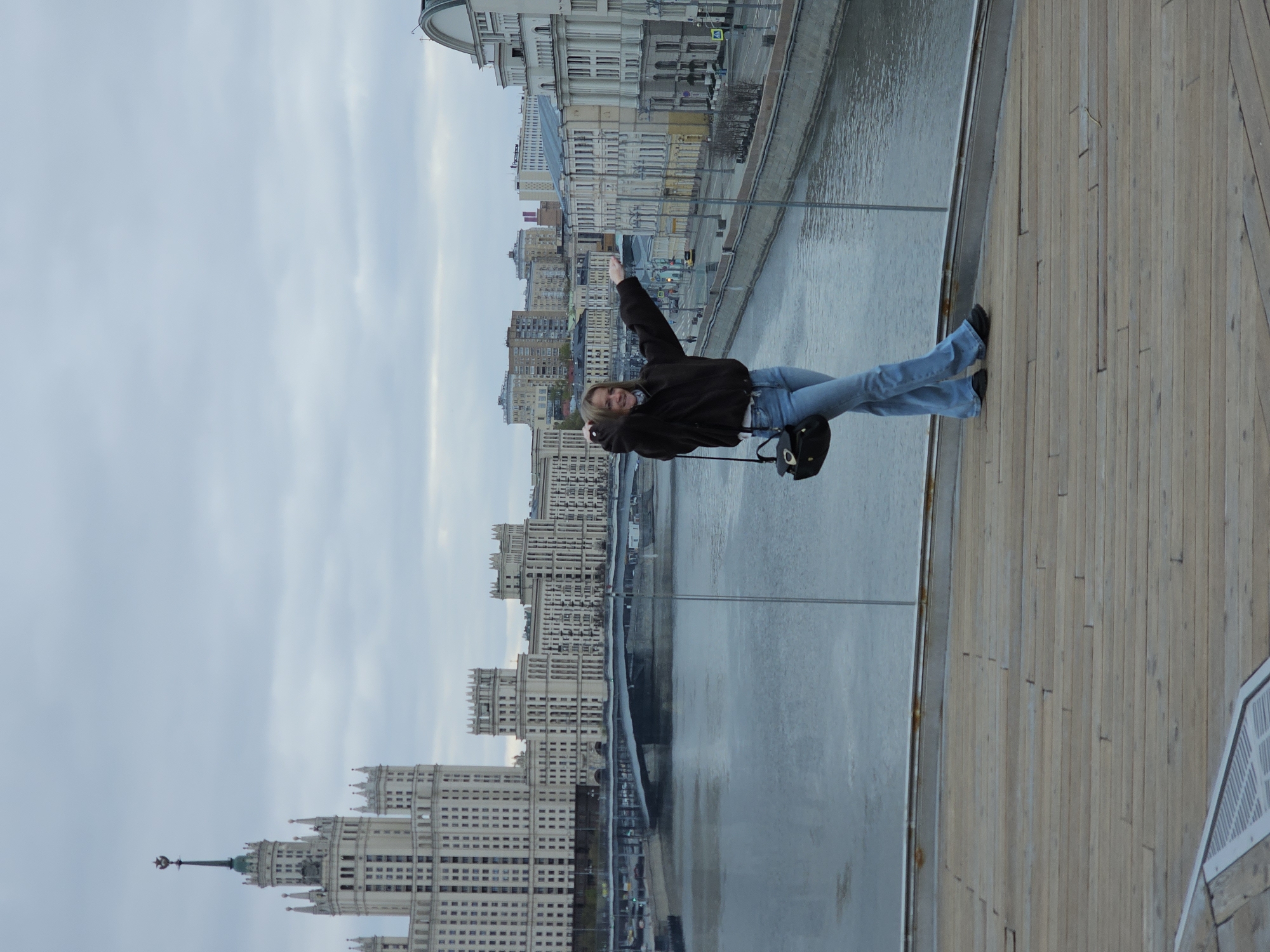 A woman is shown lying on her side in an outdoor location. She is wearing a black jacket, blue jeans, and black shoes. She is smiling and waving her right arm in the air. A black purse or bag is hanging near her.
The location appears to be a wooden boardwalk or deck adjacent to a river or body of water. Across the water, a cityscape is visible. The architecture of the buildings suggests the image was taken in Moscow, Russia. The "Seven Sisters" skyscrapers are visible, most notably the Kotelnicheskaya Embankment Building. The sky is overcast.