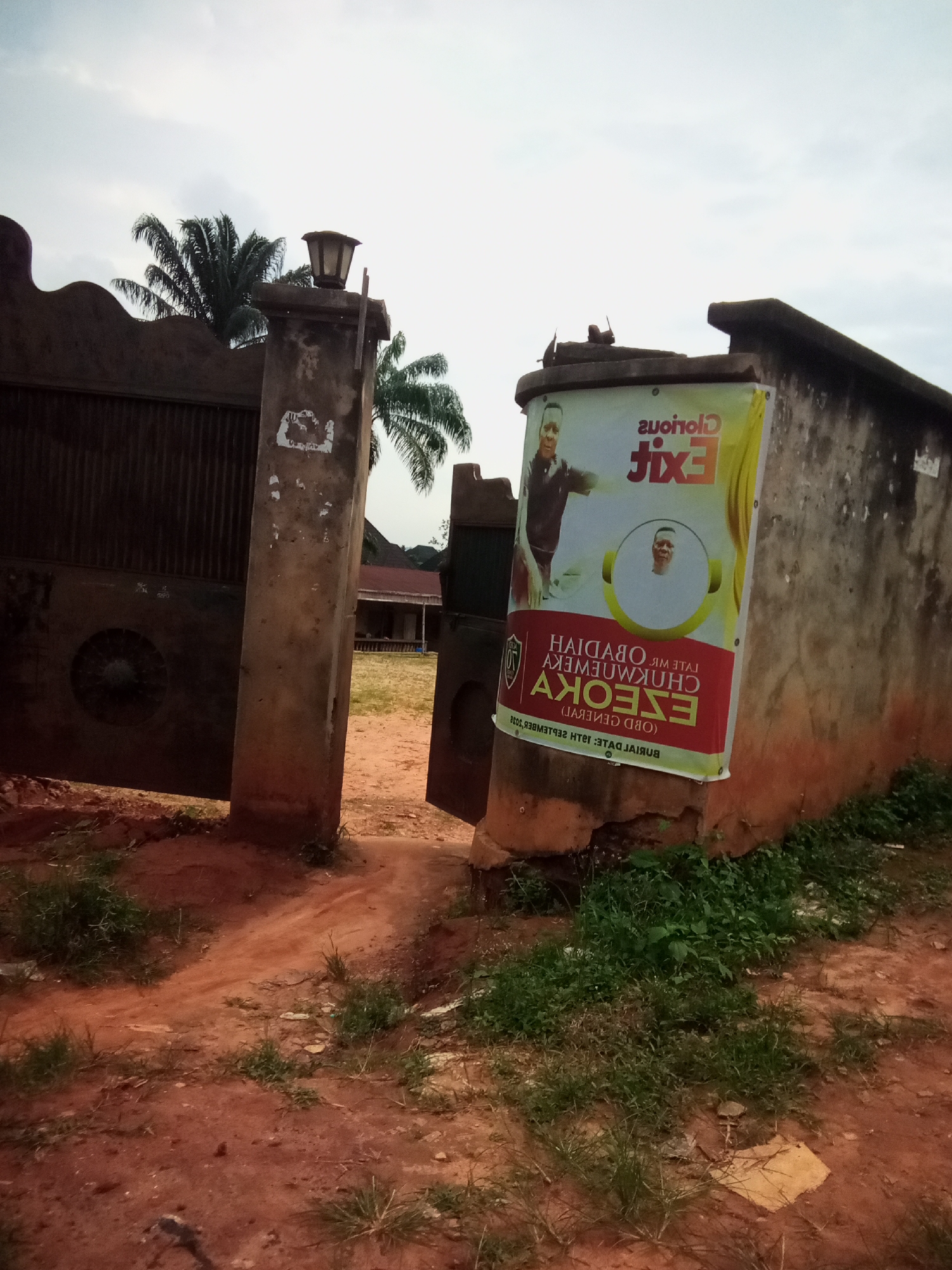 The image depicts a scene outside a property in Oko Town, Nigeria. A large, weathered metal gate, partially open, stands to the left. To the right of the gate is a concrete pillar with an attached lantern and a large banner. The banner features a man's portrait and text in what appears to be Igbo script. Behind the gate, a dirt path leads to buildings with red roofs and palm trees in the background. The ground is covered in red soil and patches of green grass. The overall impression is one of an entrance to a residential or public space.