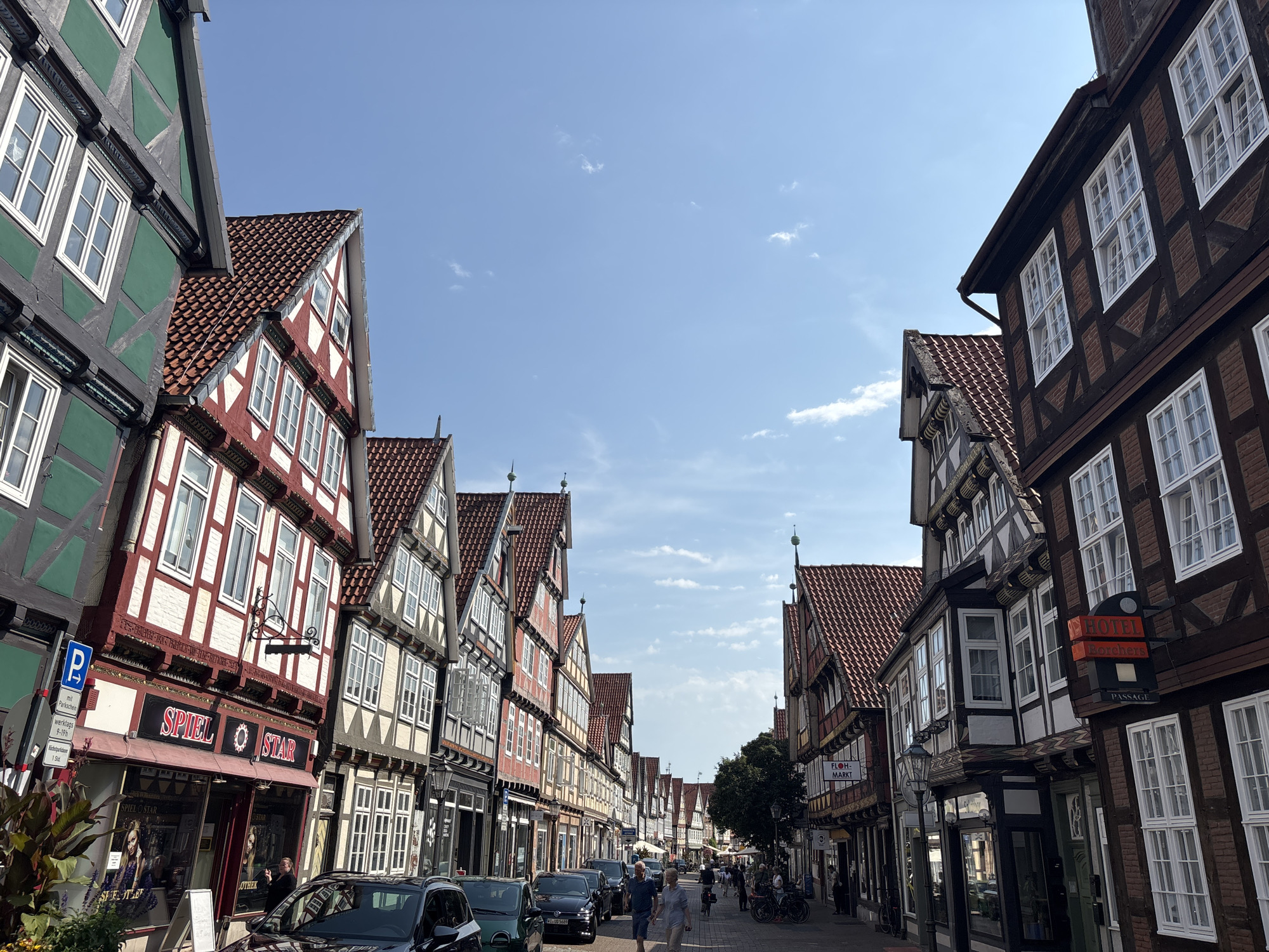 This is a street-level, eye-level, medium shot of a pedestrian street lined with historic half-timbered buildings in Celle, Germany. The buildings, with their characteristic red tile roofs and ornate facades, create a picturesque scene. The architecture suggests this is likely within the historic old town of Celle.
On the left side of the street, several buildings feature green and red facades with white trim and numerous windows. One building has a sign that reads "SPIEL STAR". A blue parking sign is visible near this building.
On the right side of the street, buildings also display the traditional half-timbered style in brown and white. One building has a sign that reads "HOTEL Borchers PASSAGE".
The street is paved with cobblestones and several people are walking along it. Some cars are parked on the street, including a black SUV and a dark green hatchback on the left. A few bicycles are also visible. The sky is blue with scattered white clouds, indicating a clear, sunny day.
The overall impression is a charming and well-preserved historic European street.