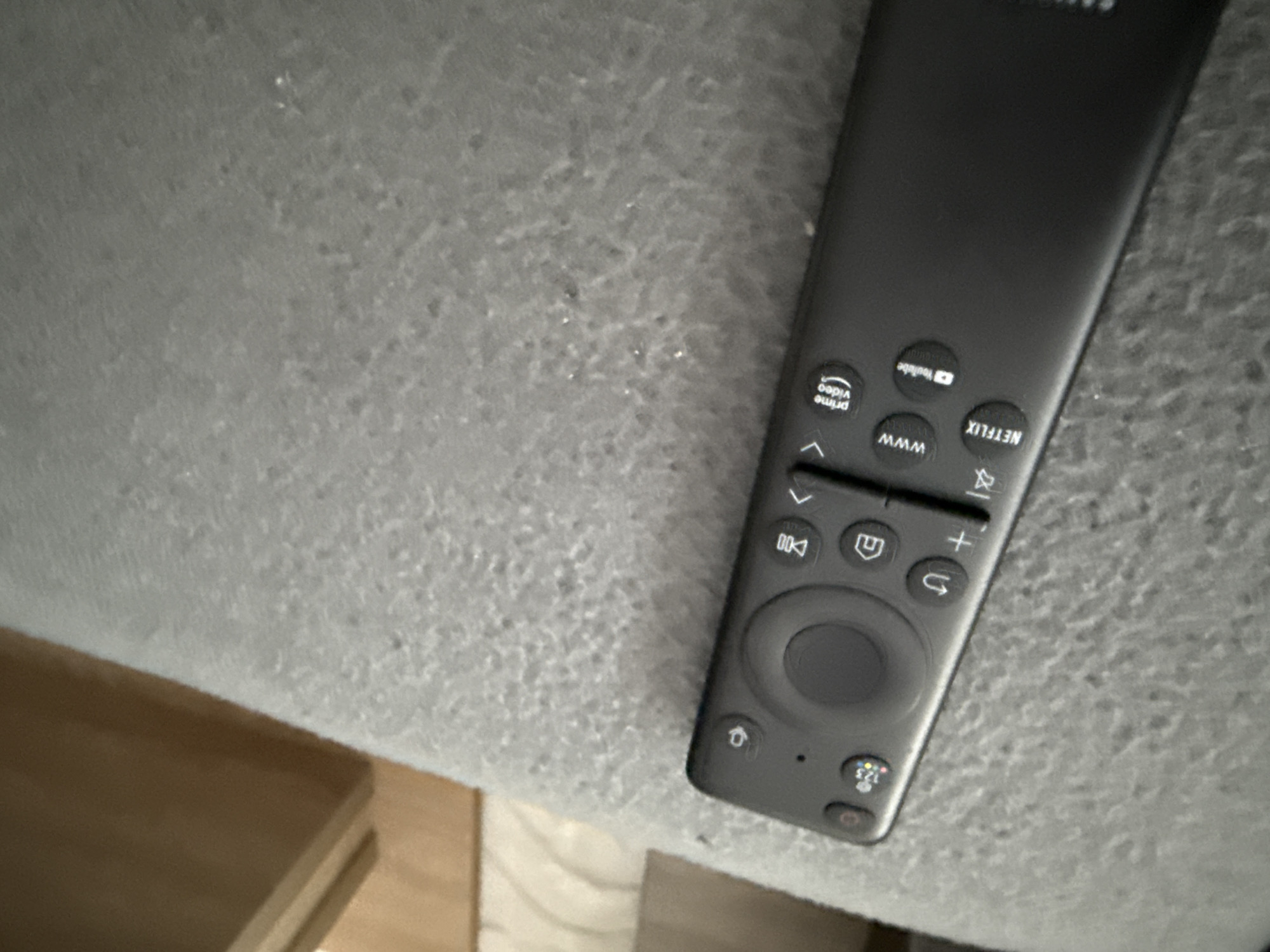 The image shows a gray Samsung remote control lying on a gray, textured surface. Part of a light brown piece of furniture is visible in the lower left corner. The remote has buttons for various streaming services, including Prime Video, YouTube, and Netflix. No information suggests this image was taken in Lovech, Bulgaria. There are no people visible in the scene.