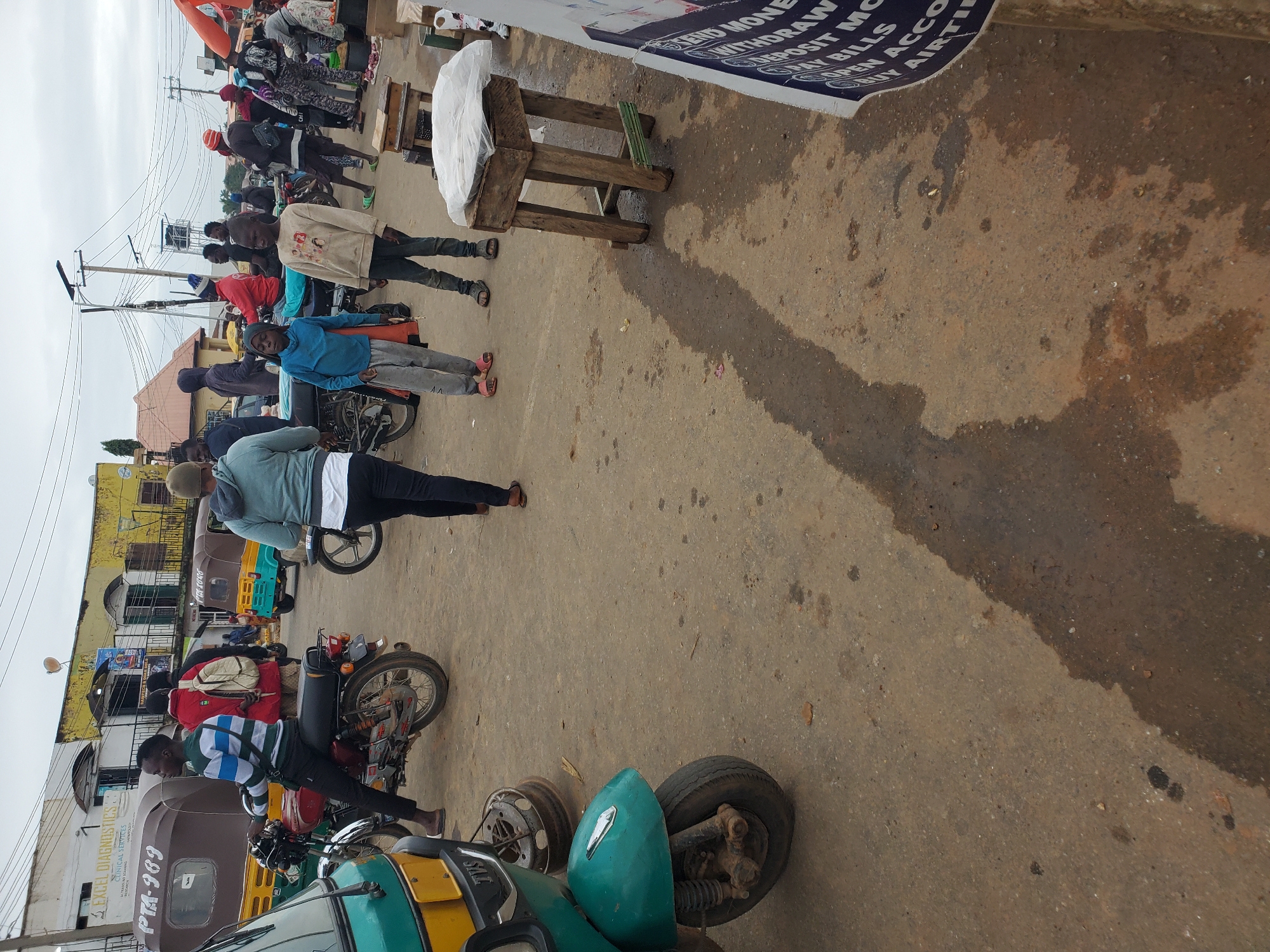 This image captures a bustling street scene in Gwong, Nigeria. The main subjects are several people, predominantly men, engaged in various activities. Some are standing, others are seated on or near motorbikes and a trishaw (auto-rickshaw), suggesting transportation and commerce. One man, wearing a blue hoodie, appears to be walking, while others are interacting with the vehicles.
The setting is a street with a worn, uneven asphalt surface, indicative of a busy commercial area. The background reveals buildings with yellow and white facades, some with visible windows and signs, hinting at shops or businesses. Overhead, power lines and poles are prominent, typical of urban environments. The weather appears overcast, with diffused light suggesting it's likely daytime, possibly morning or afternoon, but not bright sun.
Several objects are notable: a red and white motorbike is parked prominently, and a green and yellow trishaw is also visible. A wooden stall with a white-wrapped object on top is present in the foreground, possibly used for selling goods. The ground shows patches of wetness and dirt, suggesting recent rain or general street conditions.
Visible text includes "EXCEL DIAGNOSTICS CLINICAL SERVICES PTA-989" on a vehicle, indicating a local business. Another sign reads "SEND MONEY WITHDRAW DEPOSIT M BILLS OPEN ACCO BUY AIRTI," which appears to be a financial service advertisement. The overall impression is one of everyday life and economic activity in a Nigerian town. The emotions conveyed are neutral, focusing on the transactional and movement-oriented nature of the scene.