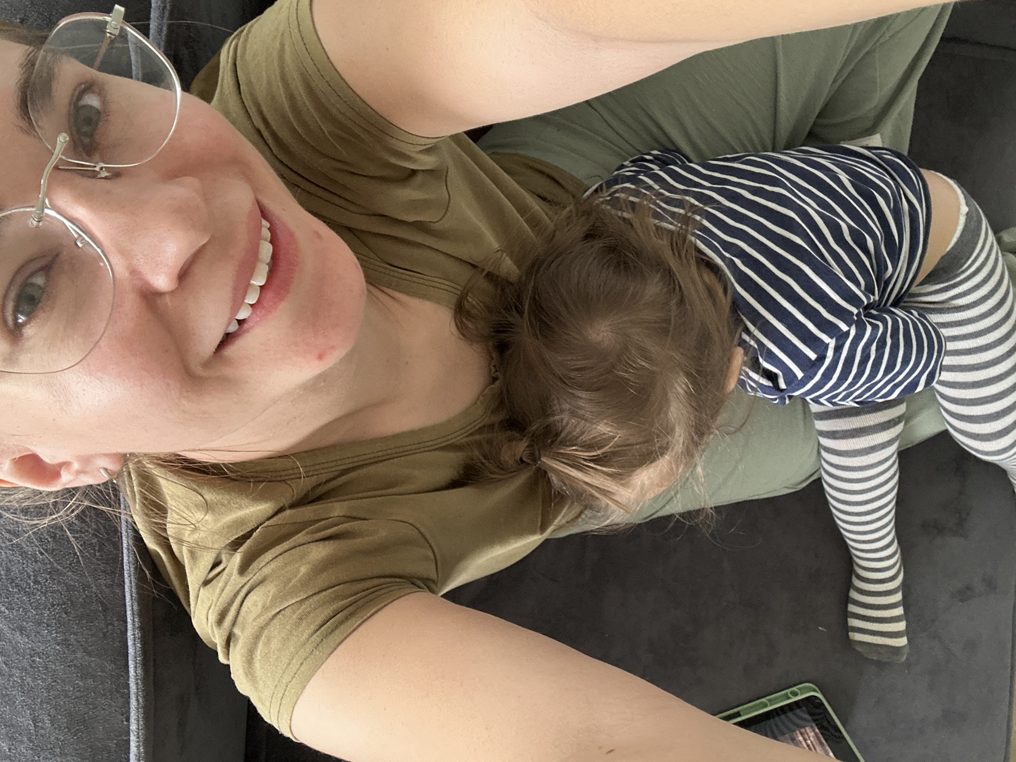 A woman wearing glasses and a green t-shirt is lying on a grey couch. A toddler is lying on top of her, wearing a blue and white striped shirt and grey and white striped leggings. A green tablet is visible in the lower right corner of the image. The location is unknown and the city of Kharnavy, Russia cannot be confirmed based on the provided image.