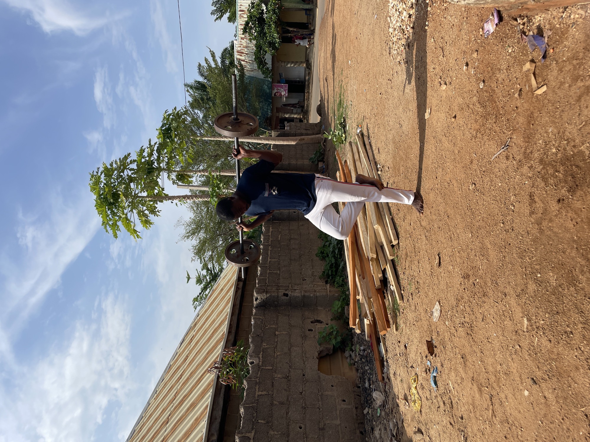 A man is lifting a barbell overhead in an outdoor setting. He is wearing a dark blue shirt and white pants and is barefoot. He is standing on a dirt ground next to a pile of wooden planks. In the background, there are buildings with corrugated metal roofs, and tall trees. The location is Akwanga, Nigeria.
