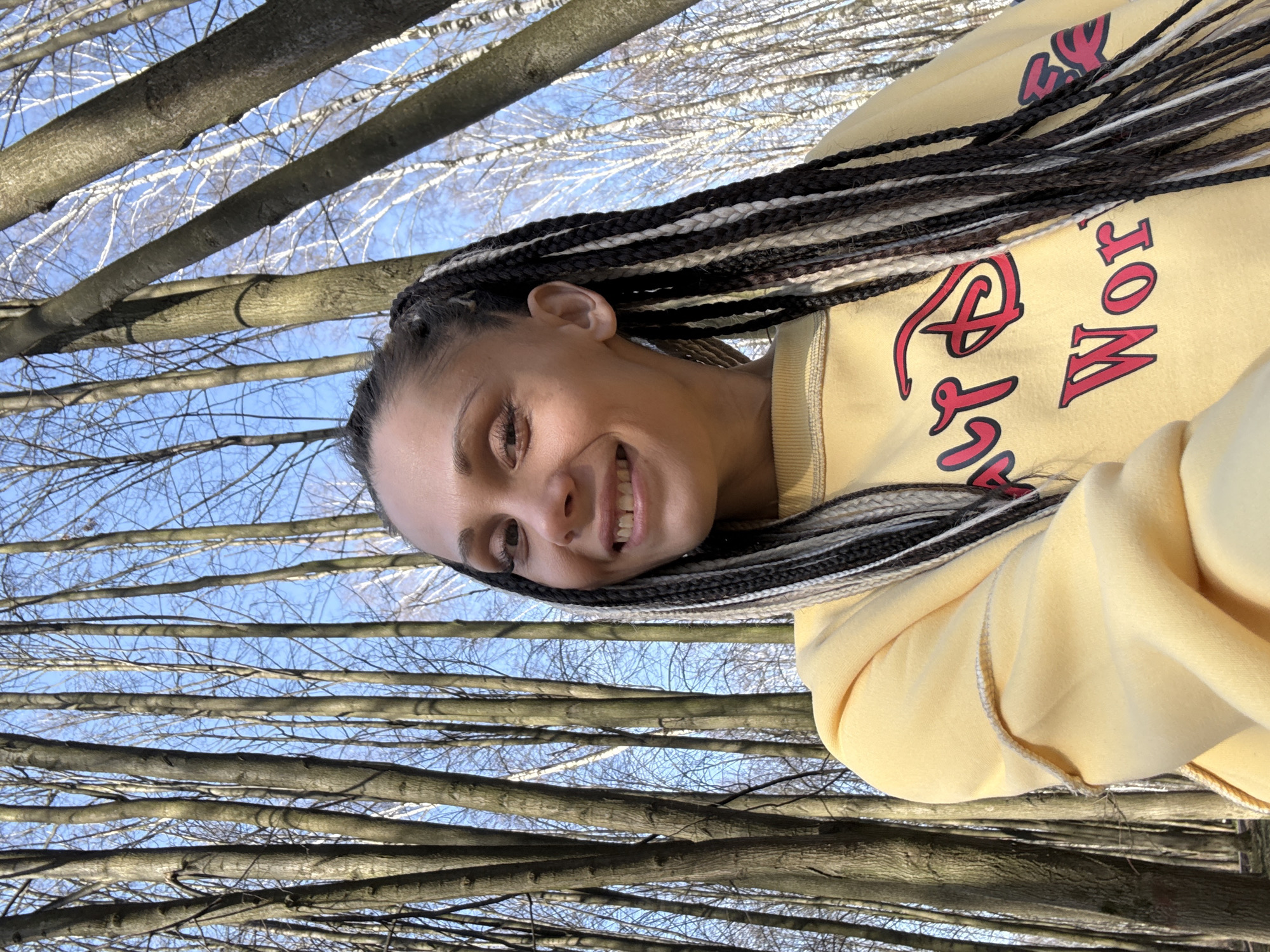 A woman with long, black and white braided hair, wearing a yellow sweatshirt with red lettering, smiles at the camera. She is in a wooded area with bare trees, likely in the city of Bor, Russia, given the prompt's context. The sky is visible through the tree branches.