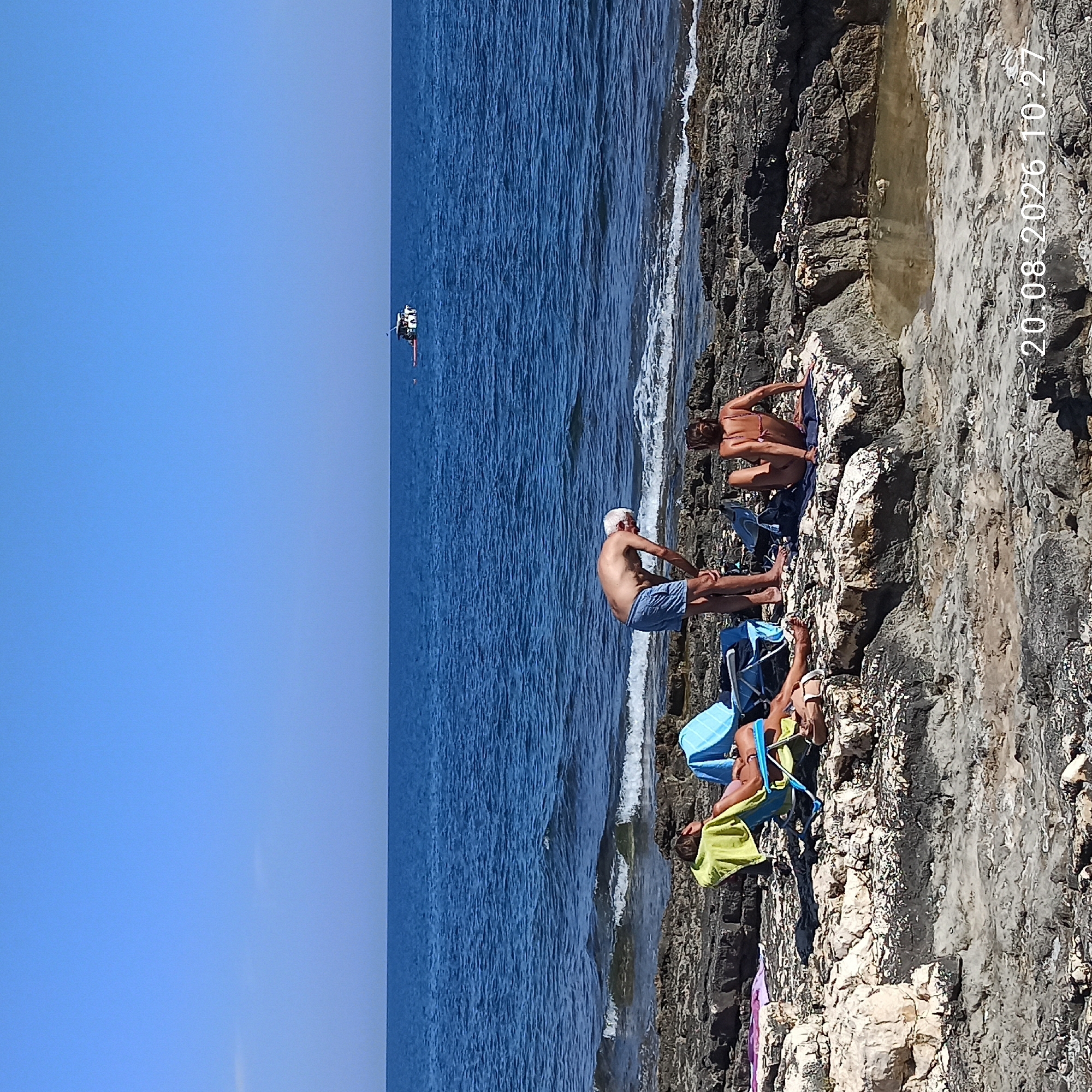 The image depicts a rocky coastline near Molfetta, Italy, on a clear, sunny day. The deep blue sea stretches to the horizon, where a small white boat with a few occupants is visible. The foreground is dominated by a rugged, dark rocky shore, interspersed with light-colored rock formations and tide pools.
On the rocks, three individuals are engaged in leisurely activities. To the left, a person lies reclined in a blue beach chair, draped with a bright green towel. Their legs are extended, and they appear relaxed. Next to them, a blue beach umbrella stands open. In the center, a man with gray hair and wearing blue shorts is bending over, possibly examining something on the ground or adjusting an object. To the right, a woman sits on a dark towel on a rock, facing away from the camera and towards the sea. She is wearing a bikini.
The overall scene conveys a relaxed summer atmosphere at the seaside. The date and time stamp "20.08.2026 10:27" is overlaid on the bottom right of the image.