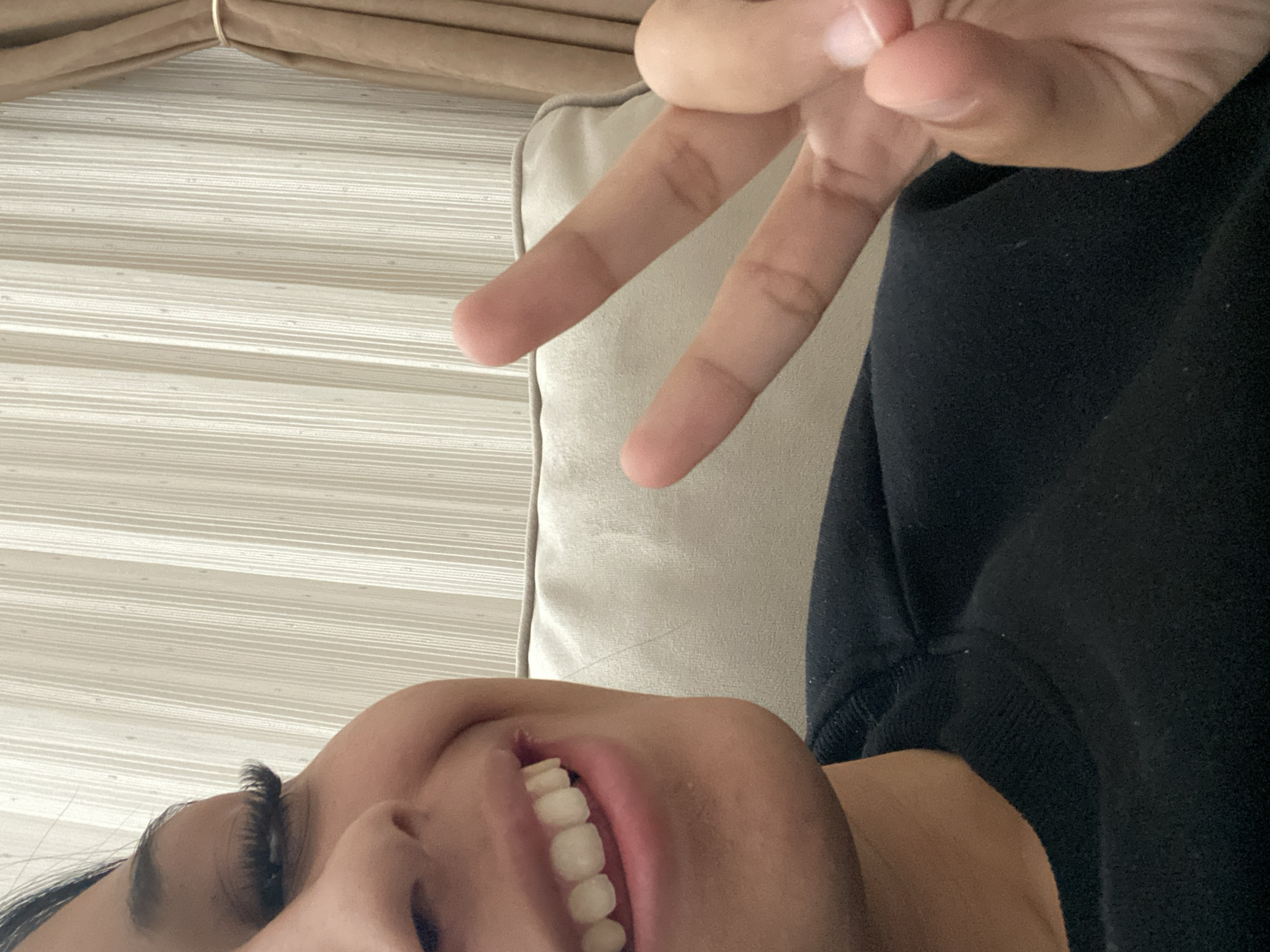 An individual, likely female, is positioned close to the camera with a partial view of their face and hand making a "V" sign. Visible features include an open mouth revealing white teeth, eyelashes, skin tone, and part of a black garment, possibly a sweatshirt. The background includes a neutral-colored, textured surface, possibly a wall or blinds, and a soft, cushioned object. The location in Saedinenie, Bulgaria is not apparent from the image.