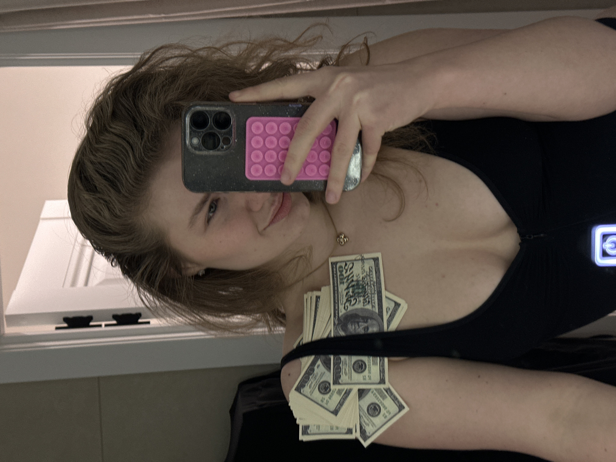 A selfie, taken in a mirror, shows a woman with wavy light brown hair. She is holding a smartphone with a pink bubble grip on the back, obscuring part of her face. She is wearing a black low-cut top. A stack of US hundred dollar bills is placed on her chest and arm. The reflection in the mirror shows a window frame. There are no indications of Kazan, Russia.