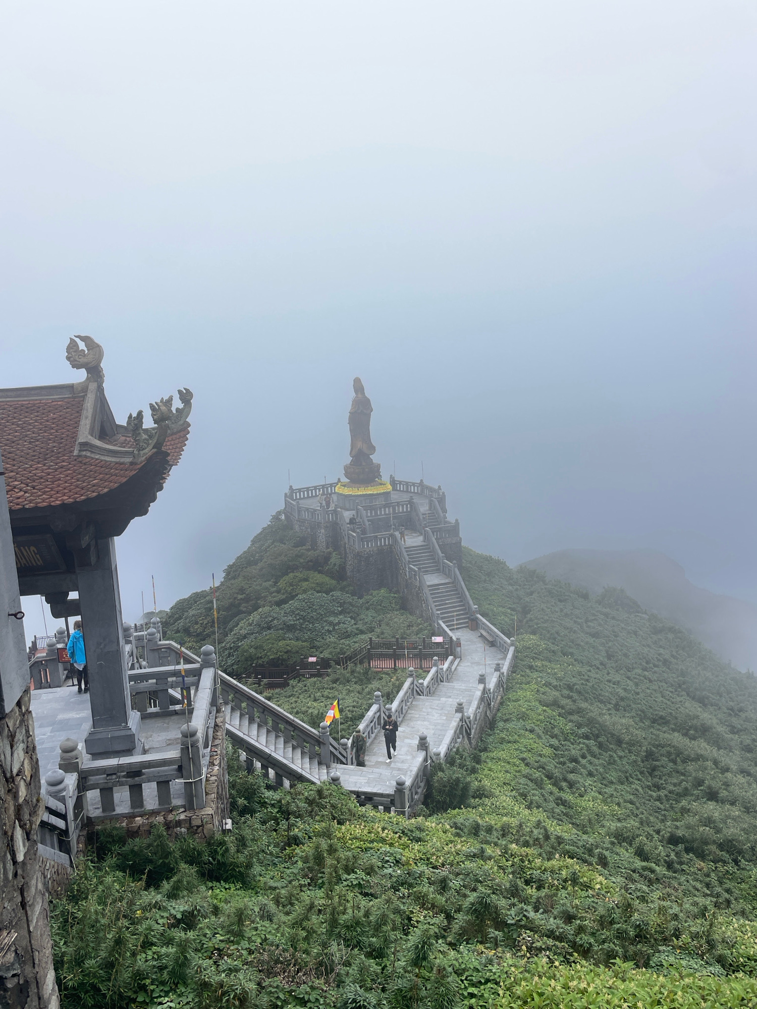 In the city of Sa Pa, Vietnam, a massive bronze statue of a standing figure, possibly a deity or historical personage, is situated atop a lush, green mountain. The statue is reached by a grand staircase with ornate railings, which winds its way up the steep incline. The architecture of the staircase and the surrounding structures suggests a religious or historical site. In the foreground, a partially visible temple-like building with a tiled roof and intricate carvings adds to the cultural ambiance. The environment is misty and overcast, partially obscuring the background and creating a dramatic, ethereal atmosphere. The vegetation is dense and verdant, carpeting the mountain slopes. Two individuals are visible on the staircase, one in a blue jacket ascending, and another in darker clothing descending. The overall scene evokes a sense of grandeur, spirituality, and natural beauty, typical of mountainous regions in Southeast Asia.