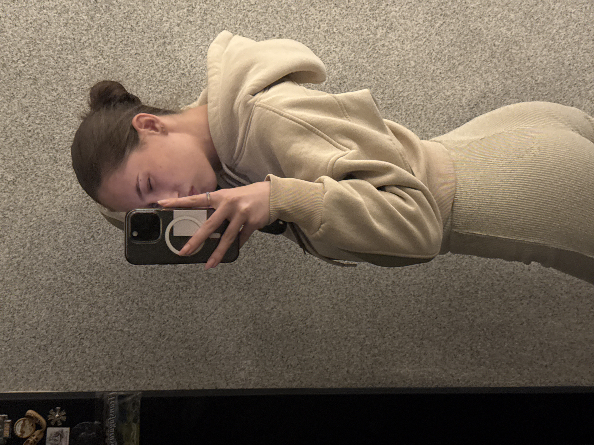 A young woman lies sideways, possibly on a bed, taking a selfie with her phone. She has dark hair pulled into a bun and is wearing a light beige hoodie and matching ribbed leggings. The background appears to be a gray carpet or wall. The bottom of the image shows a dark ledge or shelf with several small items on it. There is no indication of the city of Sofia, Bulgaria.