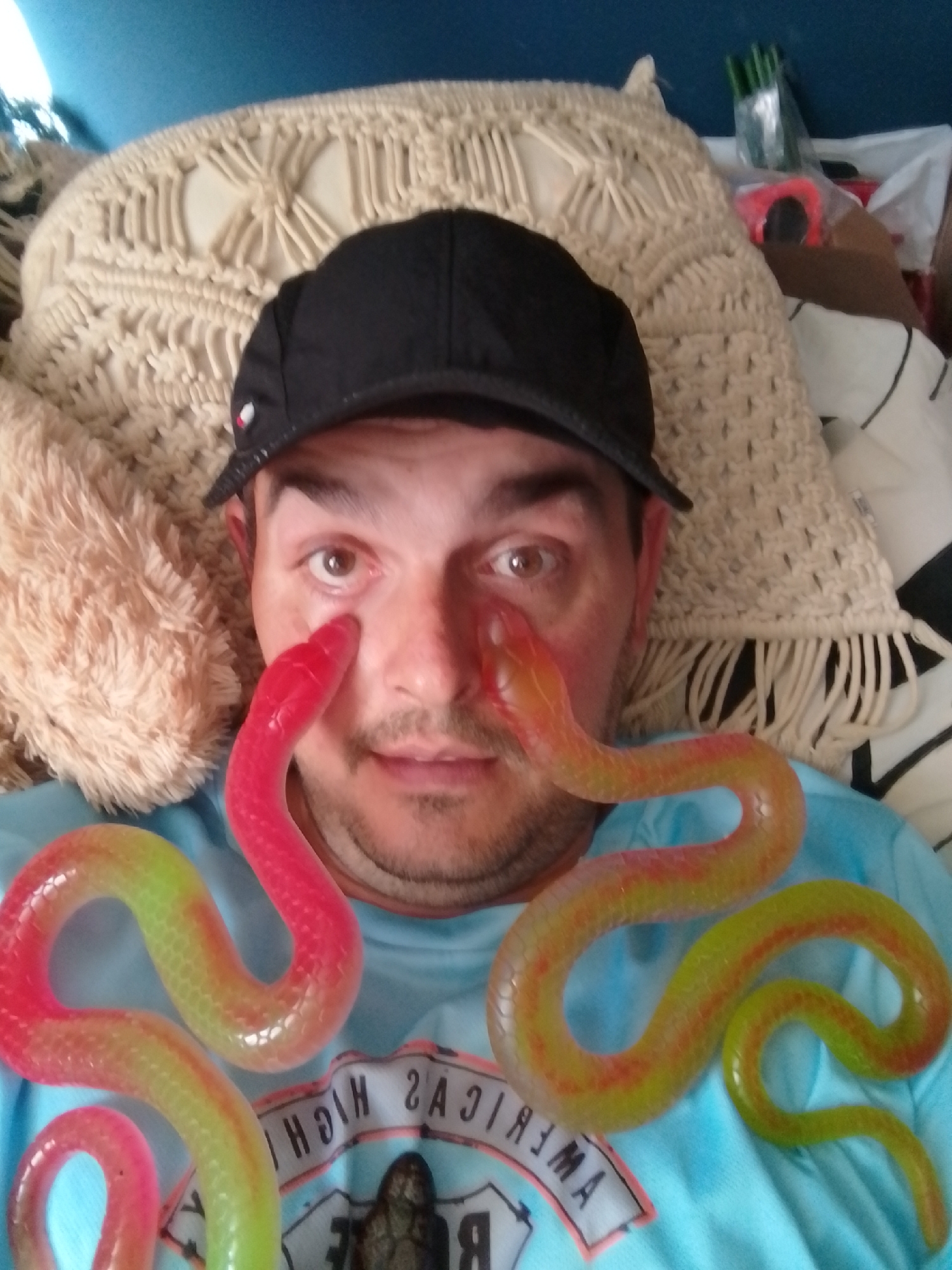 A man wearing a black baseball cap and a blue t-shirt is lying down, with his head on a cream-colored macrame pillow and a beige fuzzy pillow to his left. Two translucent, snake-shaped gummy candies, one predominantly pink and the other green and yellow, are draped across his face and shirt. One pink gummy snake extends from his left cheek towards his left eye, and a green and yellow gummy snake curves across his nose and mouth. The man's eyes are open, and he appears to be looking at the camera. The background includes a blue surface above the pillows and a white and black striped fabric behind him. The image is a selfie taken from a slightly elevated angle. There is no indication of the city of Wedel, Germany in the image.