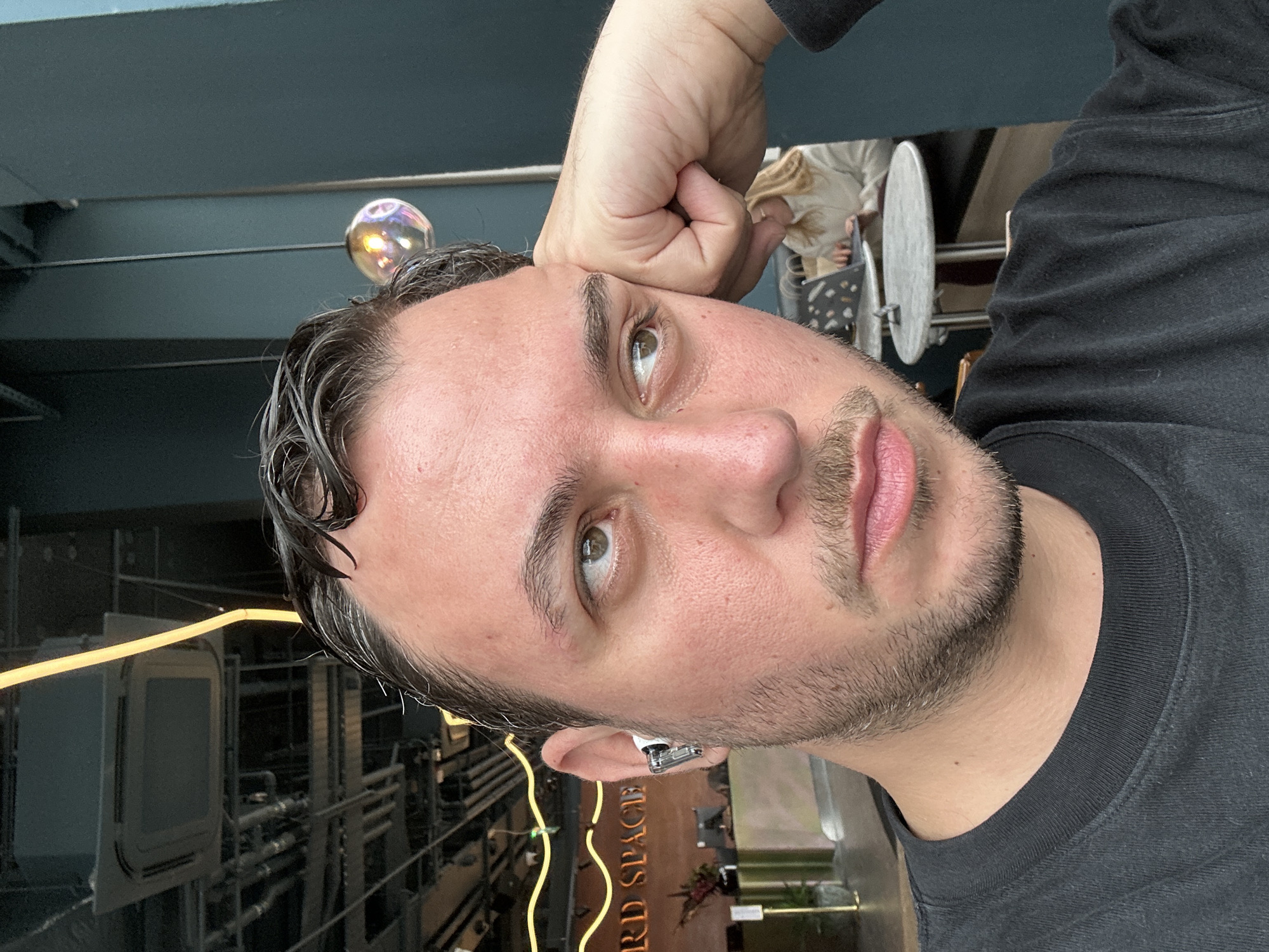 A man is posing for a selfie. He has short brown hair and a short beard. He is wearing a black long-sleeved shirt. His right hand is above his head. He appears to be wearing a white wireless earbud in his left ear. The background includes a yellow neon sign that reads "RD SPACE". A decorative soap bubble is floating in the air behind the man's head. The location appears to be a commercial interior, possibly a bar or restaurant in London, United Kingdom.