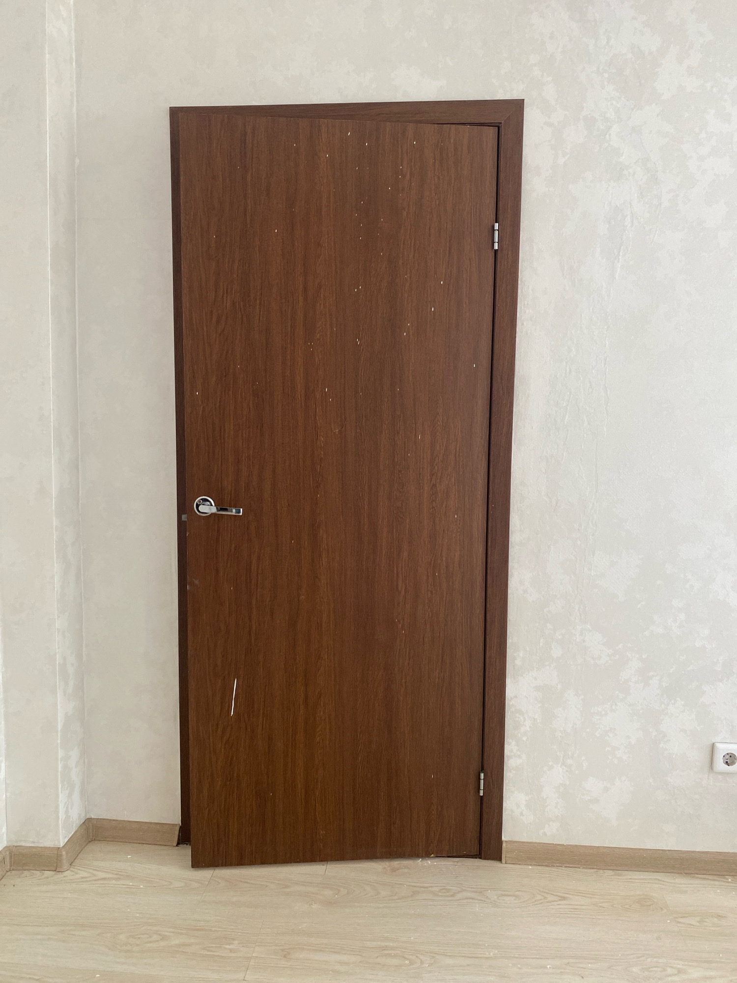 The image shows a closed wooden interior door with a silver handle, set within a white textured wall. The floor is a light-colored laminate. There is a white electrical outlet visible on the right wall. The overall scene appears to be an interior residential or commercial space. No specific location in Oyskhara, Russia is discernible from the visual information provided.