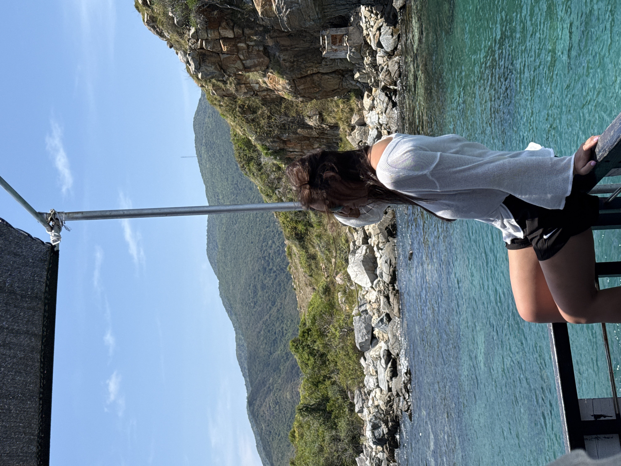 A woman leans over a railing, looking down at the water. She is wearing a white shirt and black shorts. The water is a turquoise color. Behind her is a rocky cliff with green vegetation. The sky is blue with some clouds. Based on the presence of Bac Nha Trang Ward, Vietnam, this is likely a coastal area with cliffs and clear water.