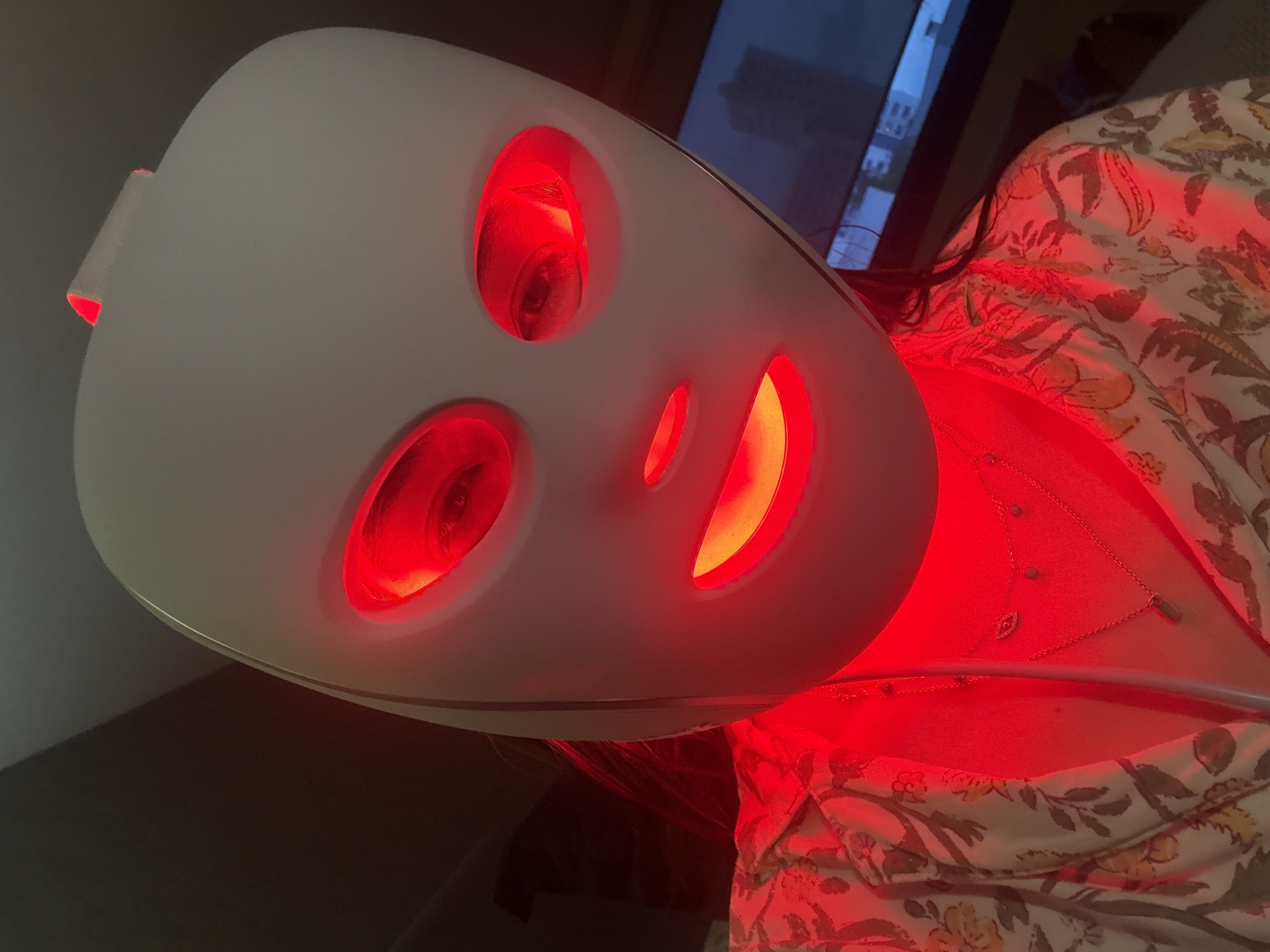 A person wears a white LED facial mask that emits red light, illuminating their face and upper chest. The mask covers most of their face, with visible eye openings showing the person's eyes. Red light also emanates from the mask's mouth opening. The person is wearing a light-colored floral patterned shirt and delicate necklaces. The background is dark and out of focus, with a hint of a television screen displaying muted colors. This scene is taking place in the city of San Bernardino Tlaxcalancingo, Mexico.