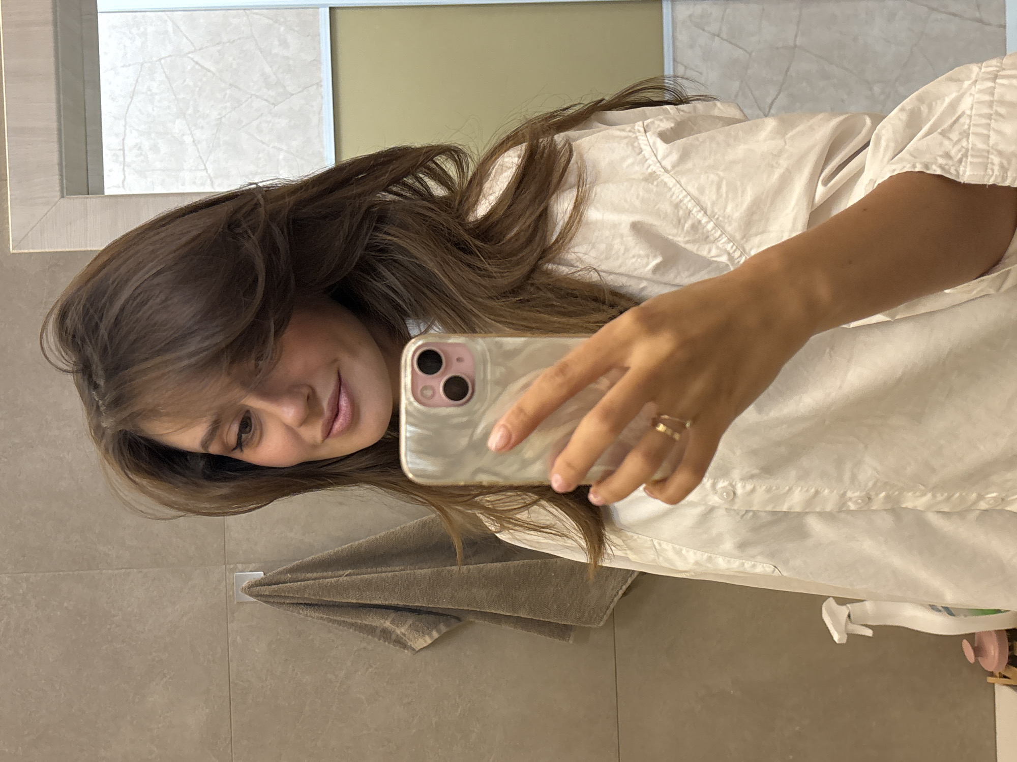A young woman with shoulder-length brown hair takes a selfie in what appears to be a bathroom. She is wearing a white button-down shirt. She holds a phone with a light-colored case. Her hand has a gold ring on one of the fingers. Behind her are neutral-toned tiles, a towel hanging on a hook, and a mirror showing wall panels of different textures. The location is not discernable from the image and cannot be determined to be in Krasnoyarsk, Russia.
