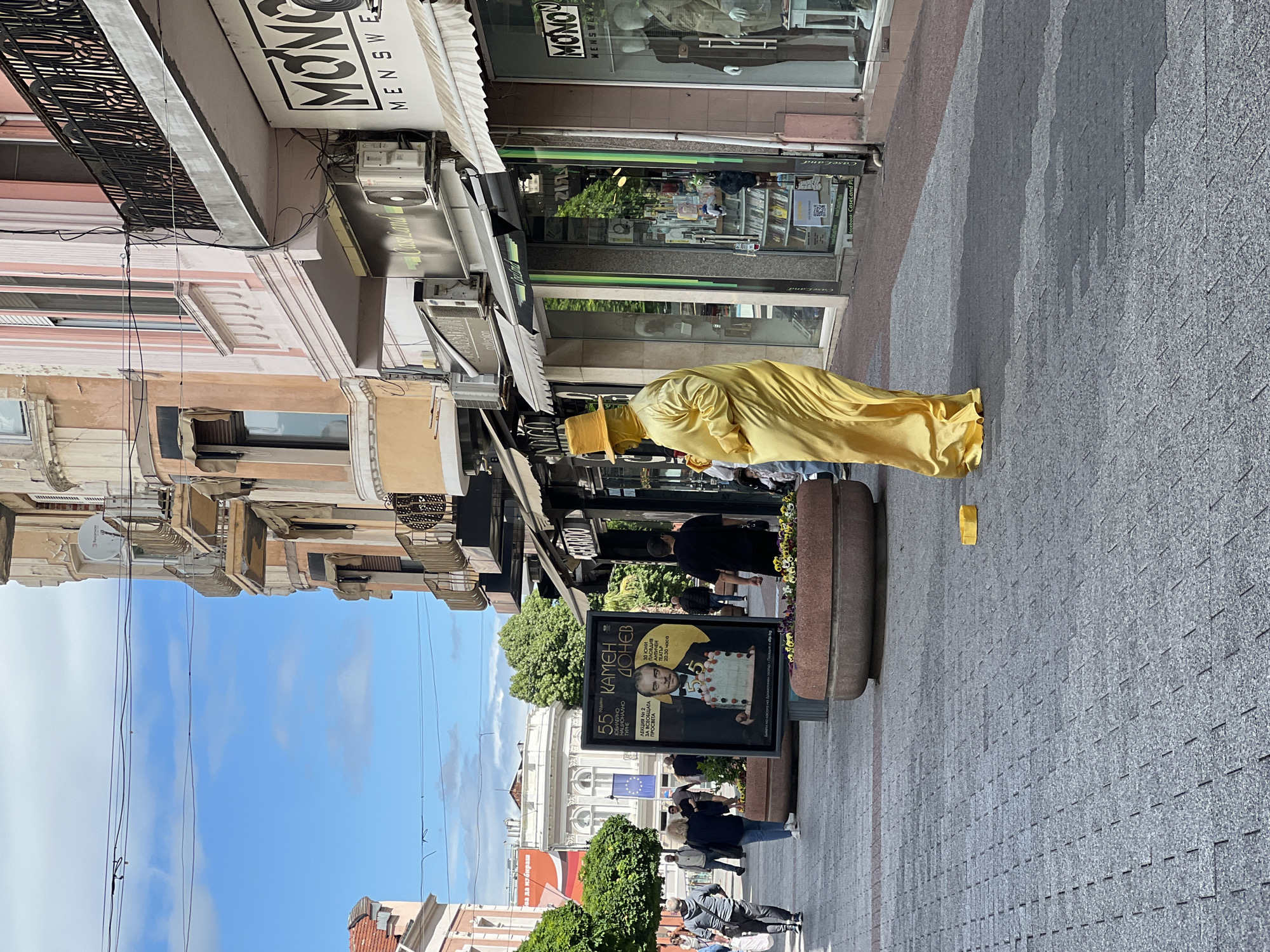 The image shows a street scene in Plovdiv, Bulgaria, on a sunny day. A person dressed entirely in yellow, including a hat, is standing on the paved sidewalk next to a flower-filled planter. Shops line the street with signs in Cyrillic script. A poster for "KAMEN DONEV" can be seen next to the planter. People are walking in the background. Buildings with ornate facades are visible across the street.