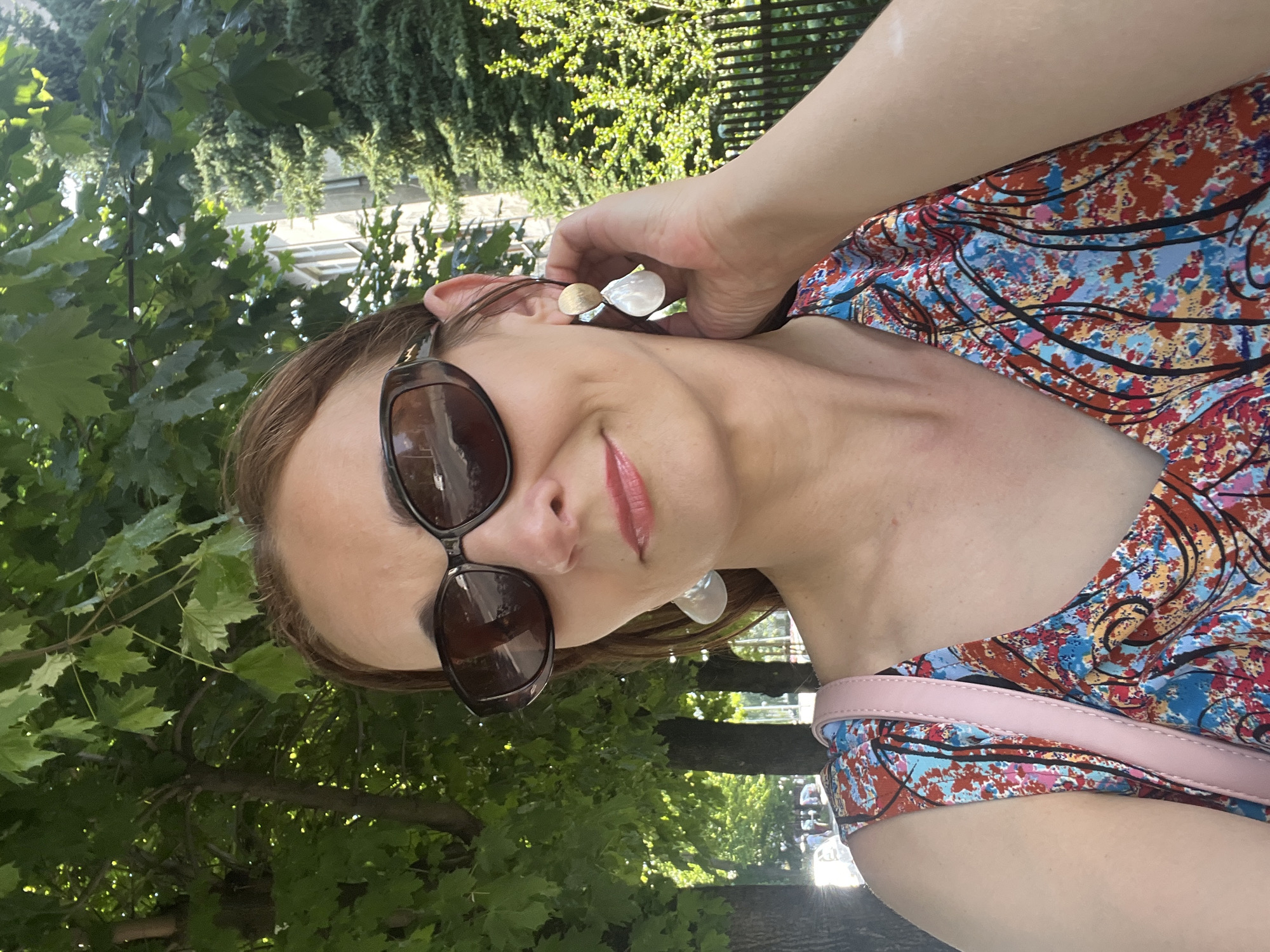 A woman with auburn hair is taking a selfie outdoors, with trees and a building in the background. She is wearing large, dark sunglasses, a colorful dress with a red, blue, and yellow abstract pattern, and large, light-colored dangling earrings. A pink strap from a bag is visible across her shoulder. Her right hand is touching her ear, near her earring. The scene appears to be in a park or along a tree-lined street on a sunny day.