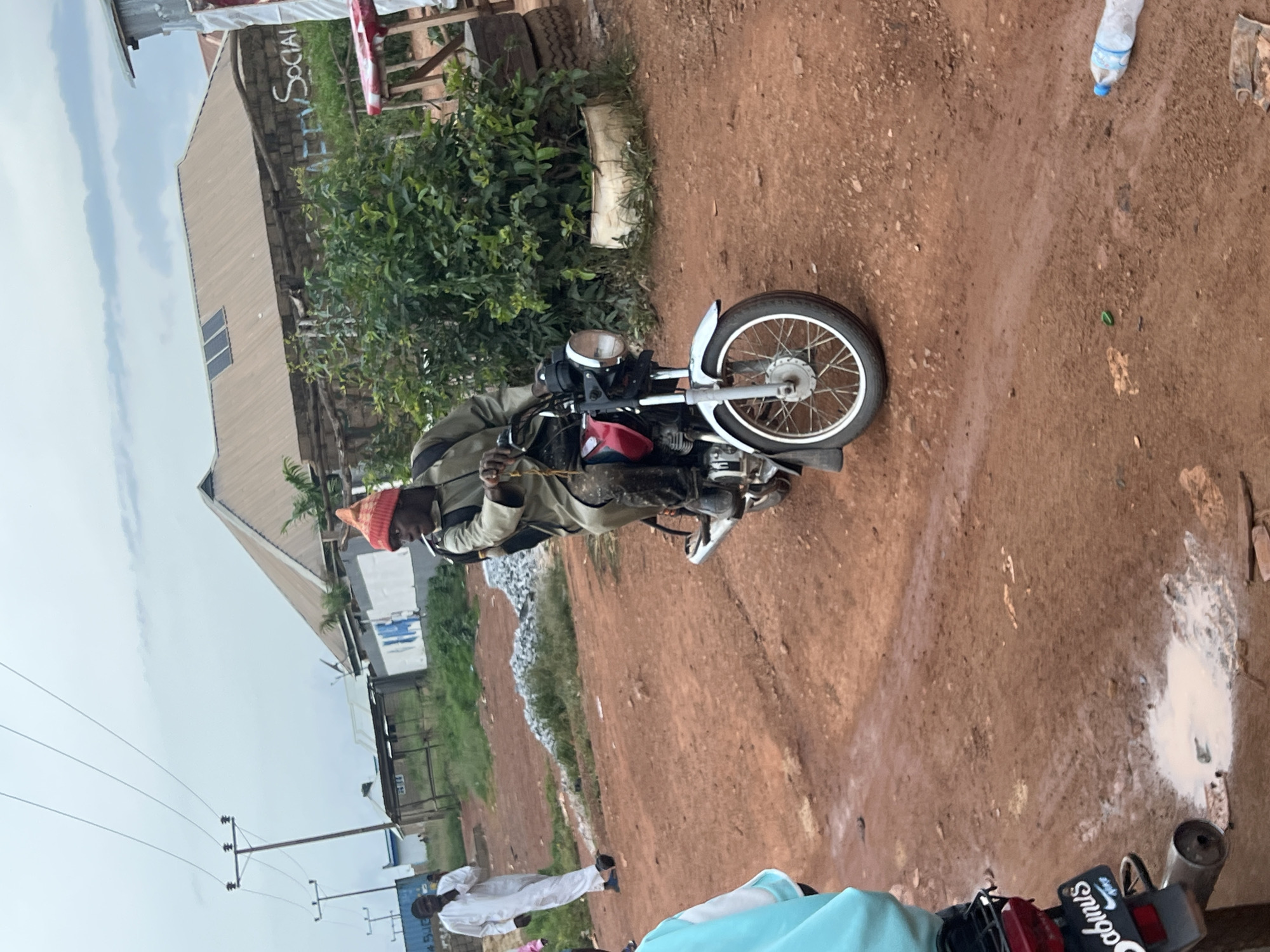 In Durumi, Nigeria, a man wearing a red beanie and a tan jacket rides a red and silver motorcycle on a dirt road. The road is muddy in places, with puddles and scattered debris. In the background, there are several buildings, some with corrugated metal roofs, and utility poles with power lines. A few other people are visible in the background, including one man in a white robe and another person in a light blue jacket. There are also some bushes and plants growing in planters made from old tires. A plastic water bottle lies on the ground near the motorcycle. The sky is overcast.