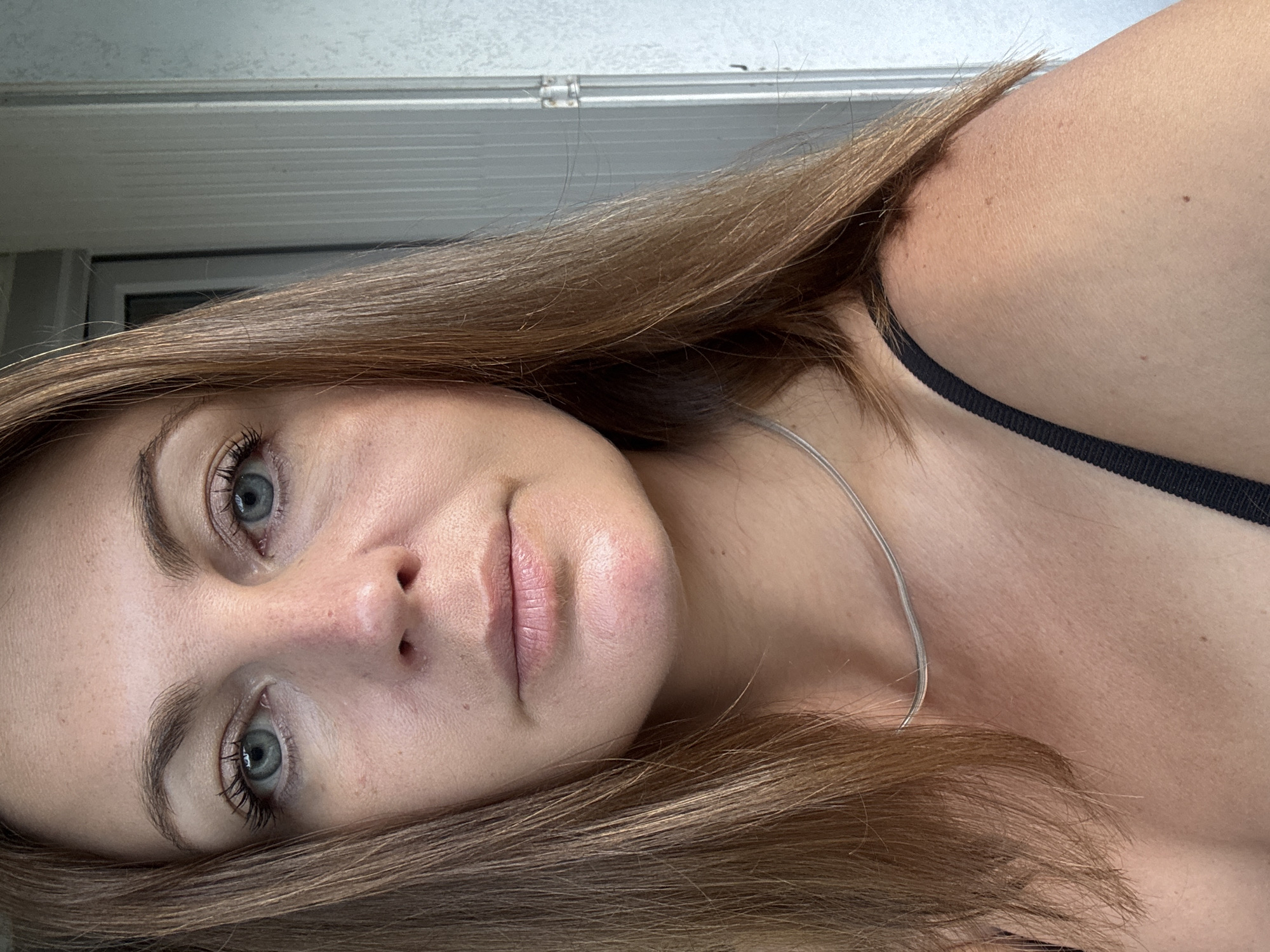 A close-up, head-and-shoulders portrait of a Caucasian woman with shoulder-length brown hair and blue eyes. She is wearing a black strap top and a thin silver necklace. The background is out of focus, showing vertical blinds or siding. The image appears to be a selfie taken outdoors, possibly on a porch or balcony. There are no discernible landmarks that would suggest a specific location within Saint-Claude, France.