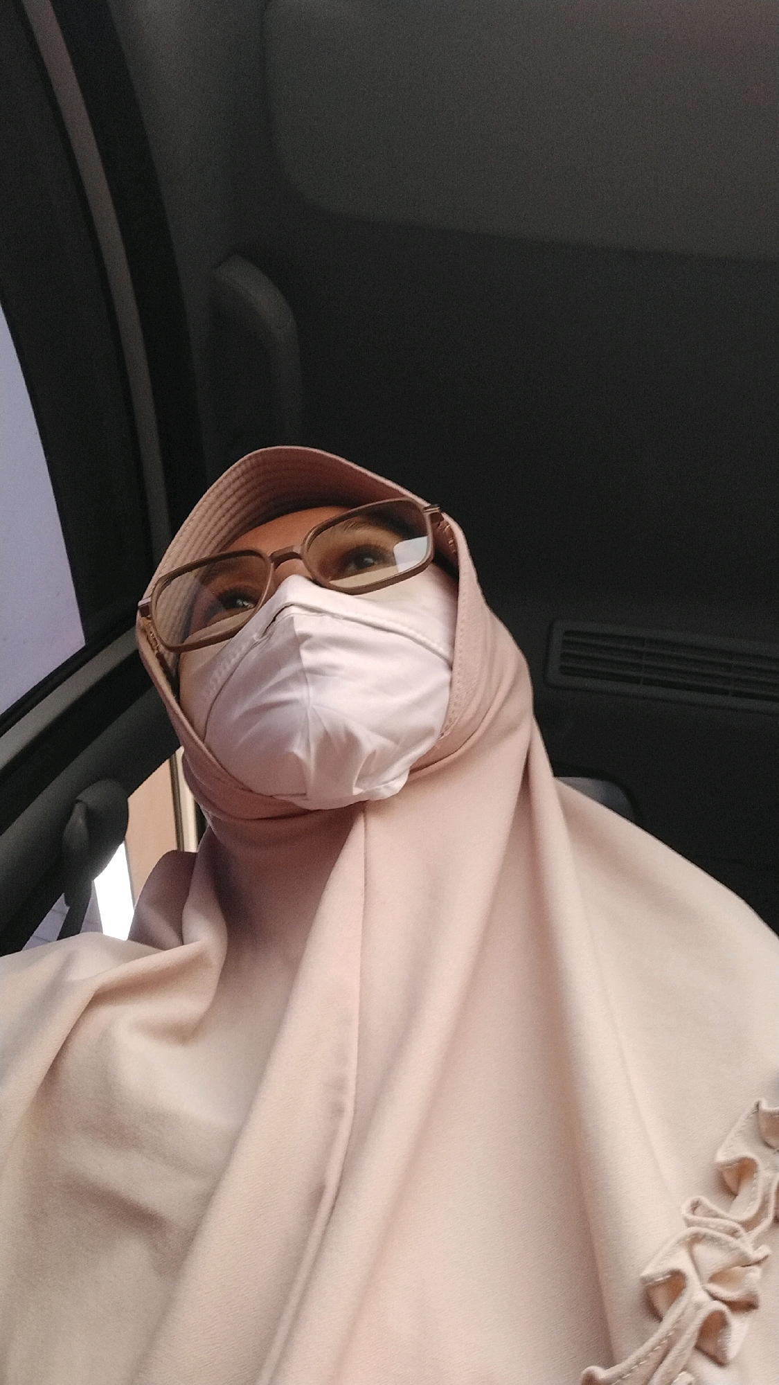 A woman wearing a light pink hijab and a light pink face mask is seated inside a vehicle. She is also wearing eyeglasses with brown frames. The interior of the vehicle is visible, including the dark-colored ceiling and a black air vent on the right side. The image is a close-up, taken from a low angle, focusing on the woman's face and upper body. The background suggests the woman is in Megang Sakti II, Indonesia.