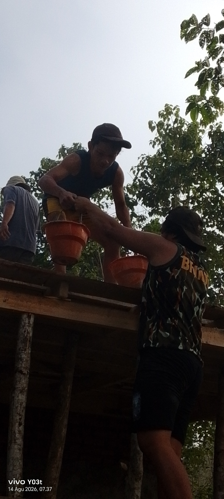 The image captures three individuals engaged in an outdoor activity, possibly construction or maintenance, on a wooden structure under a bright, overcast sky. The foreground features a person wearing a camouflage-patterned sleeveless shirt with the letters "BNN" visible on the back, and dark shorts, positioned on the right side of the frame, looking down. In the middle ground, another individual, wearing a dark sleeveless shirt and yellow shorts, is bending over, holding an orange bucket. To the left, a third person, wearing a blue shirt and a hat, is partially visible, also on the wooden structure. The structure itself appears to be under construction or in disrepair, with visible wooden beams and supports. Foliage from trees is present in the background. The timestamp "vivo YO3t 14 Agu 2026, 07.37" is superimposed on the lower left corner of the image. The scene is set outdoors, with the sky being a prominent feature, and the surrounding environment suggests a rural or semi-rural location within Lara, Indonesia.