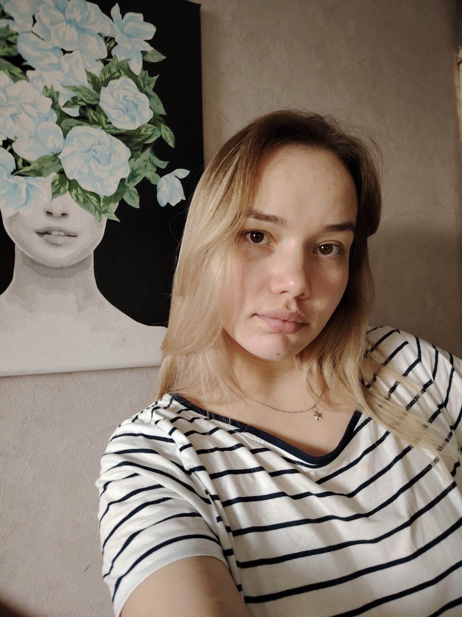 A young woman with blonde hair is pictured indoors in a close-up selfie. She is wearing a white and black striped V-neck t-shirt and a silver necklace with a star pendant. Behind her on the wall is a painting depicting a woman's face obscured by a bouquet of light blue flowers against a black background. The wall is a light beige color. The photo was taken in the city of Марковский, Russia.