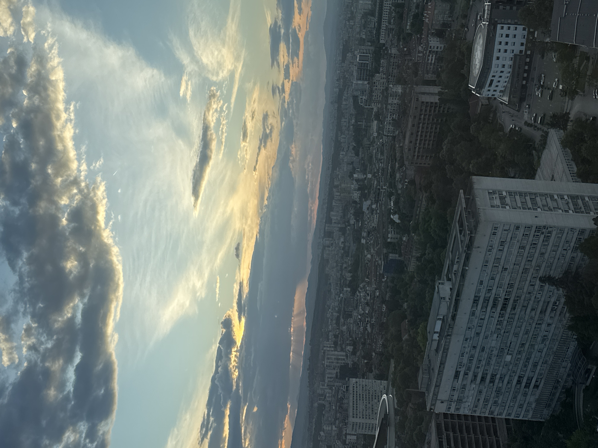 A high-angle, rotated view of Sofia, Bulgaria, shows a cityscape with buildings and trees under a dramatic sky. The left portion of the image is dominated by clouds, some illuminated with the colors of sunset. The city, comprised of dense buildings and interspersed greenery, extends towards the horizon. Several large buildings are visible, with one prominent white building in the lower right and another with a rounded edge in the upper right. Cars are sparsely scattered on the roads around the buildings. The horizon shows a light band suggesting a mountain range obscured by haze.