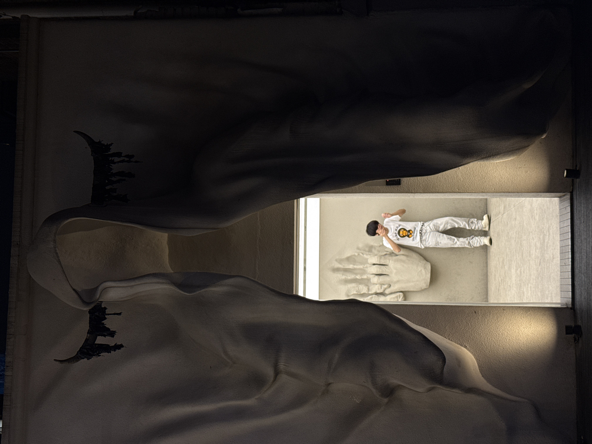 A long, slightly rotated overhead shot shows an installation. The installation features what appears to be a large sculpture that includes a pair of large, wing-like forms, one dark, and one light. On the left side of each wing-like form is a dark, jagged, abstract shape. Between the two forms is a rectangular cut-out. In the cut-out, a person wearing a white outfit lies on their back on a sculpture of an outstretched hand. There are two lights on the right side of the frame that illuminate the wall surrounding the cut-out. The location is Tianhe District, China.