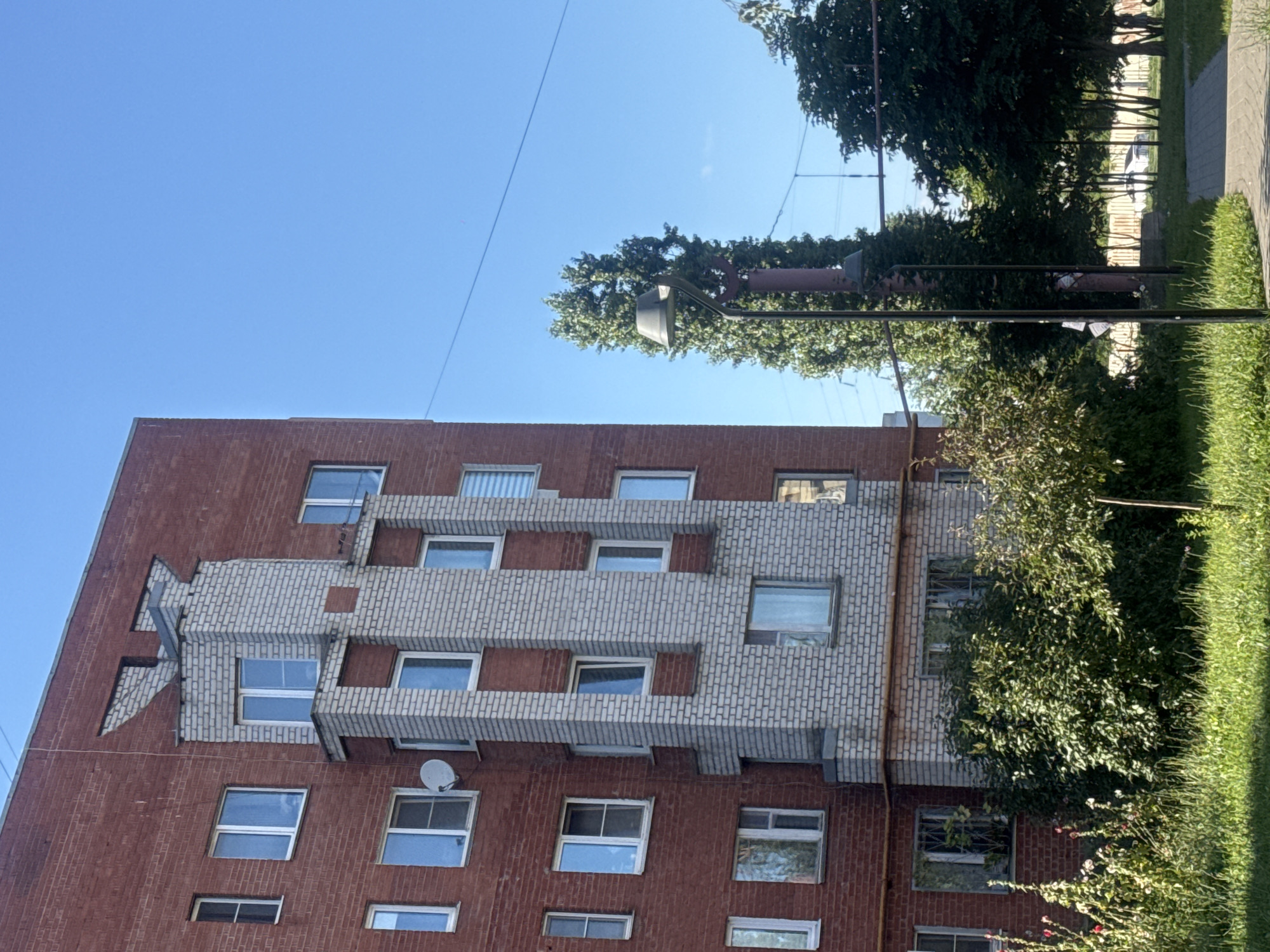 The image shows a multi-story apartment building constructed with red brick and white brick accents, featuring numerous windows. A white satellite dish is visible on the red brick section of the building. In front of the building, there is lush green grass, bushes, and some flowering plants. A paved walkway is present, and a lamppost stands near it. Tall trees are visible behind the building, with some power lines running across the clear blue sky. The scene appears to be a residential area, likely in Nizhny Novgorod, Russia, given the architectural style typical of Soviet-era apartment blocks. There are no people or active interactions depicted in the image.