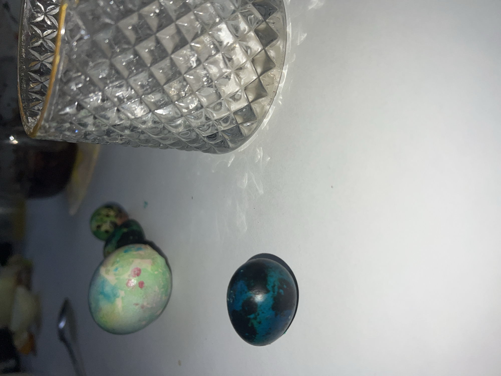 A high-angle shot depicts a crystal glass with a gold rim in the upper left corner. Three painted eggs are visible on a light-colored surface, one near the bottom left, and another at the bottom. The photo was taken indoors. The specific location in the city of Noviy, Russia, is not discernible from the image. There is no evidence of people or actions being performed.