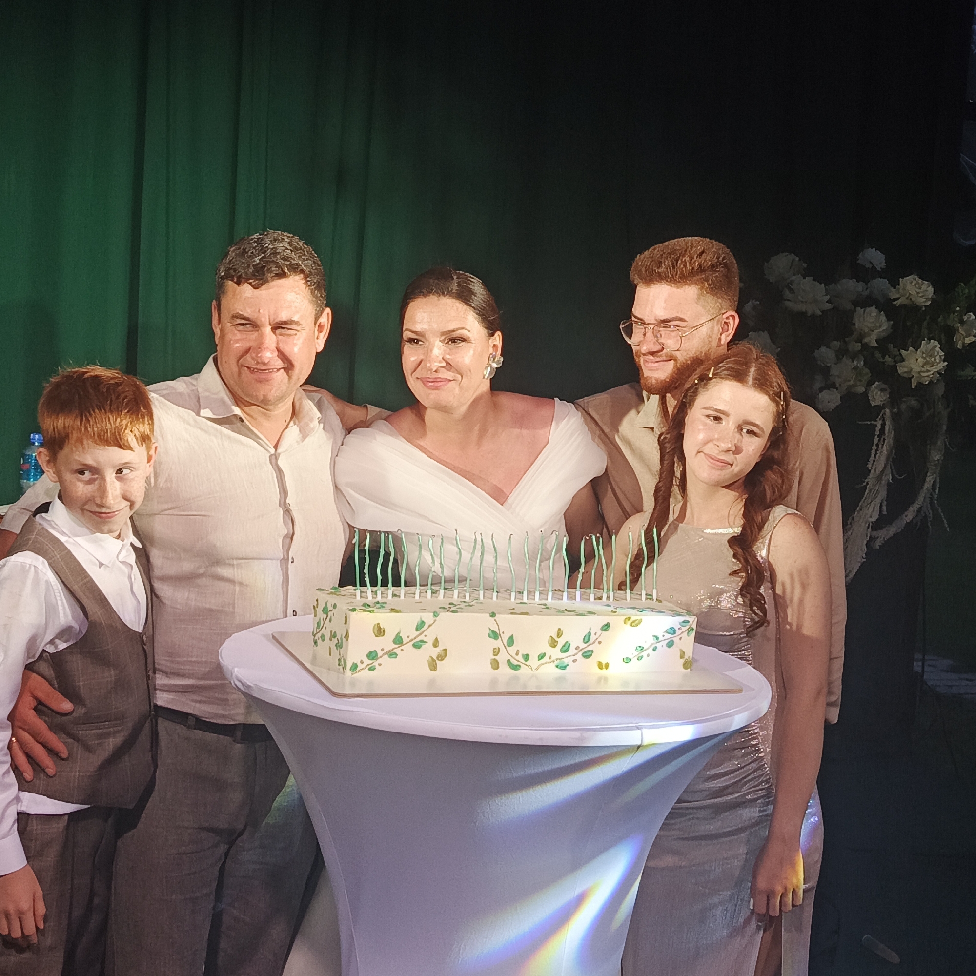 A family of five is gathered around a cake on a table, with a dark green curtain in the background and white flowers to the right. The family consists of a man on the left with his arm around a young boy with red hair. Next to him is a woman in a white dress, and to her right is a man wearing glasses. On the far right is a young girl with braids. The cake is rectangular with white frosting decorated with green and gold leaves and has multiple green candles. The lighting suggests an indoor event, possibly a celebration. The location is identified as the city of Svetloe, Russia.