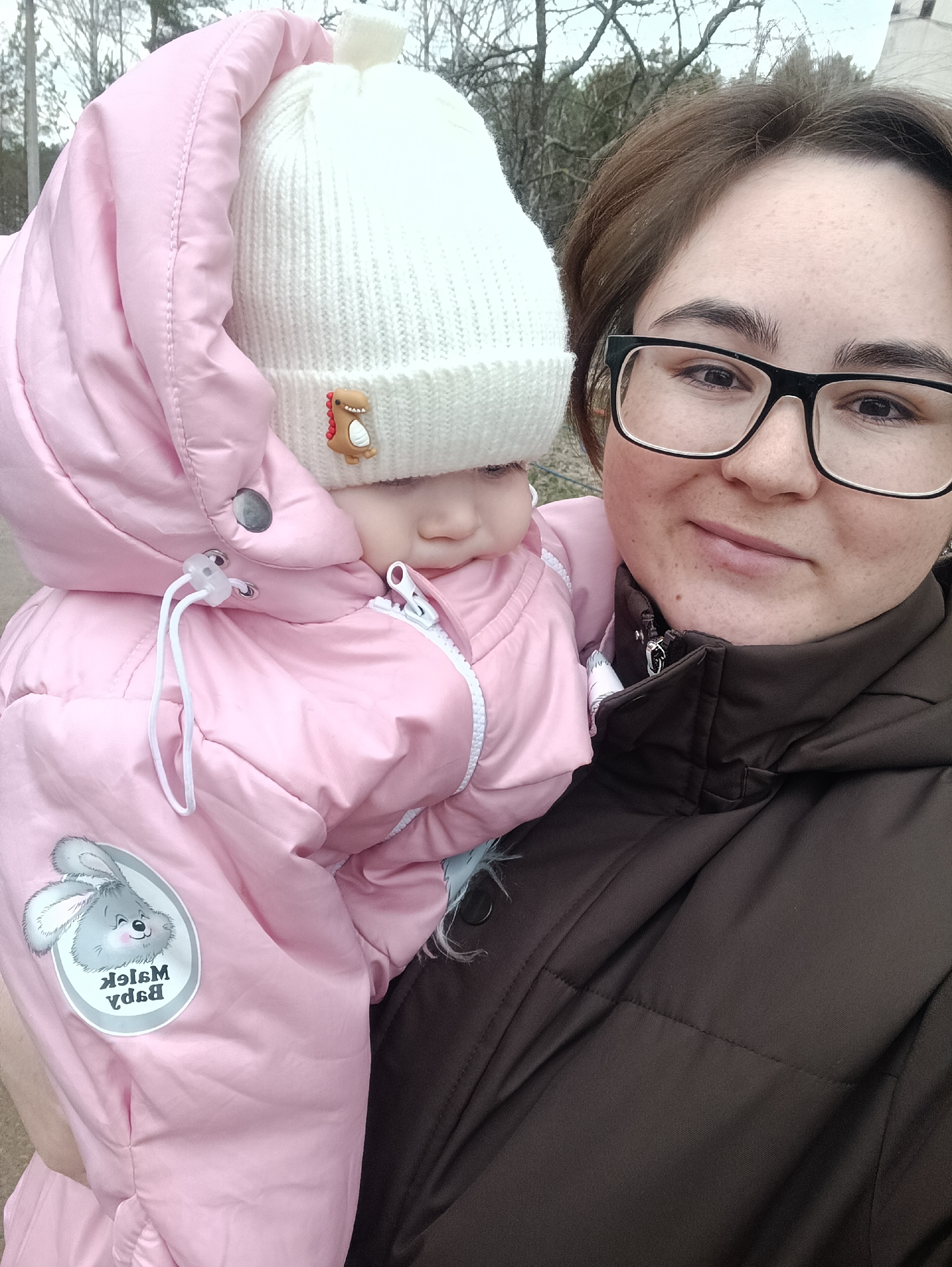 A woman in eyeglasses and a brown jacket is holding a baby wearing a pink snowsuit with a white knitted hat with a dinosaur patch. The snowsuit has a patch with a cartoon rabbit and the upside-down text "Baby Malek." The background features trees without leaves. The location is not explicitly identified as Noviy, Russia, in the image.