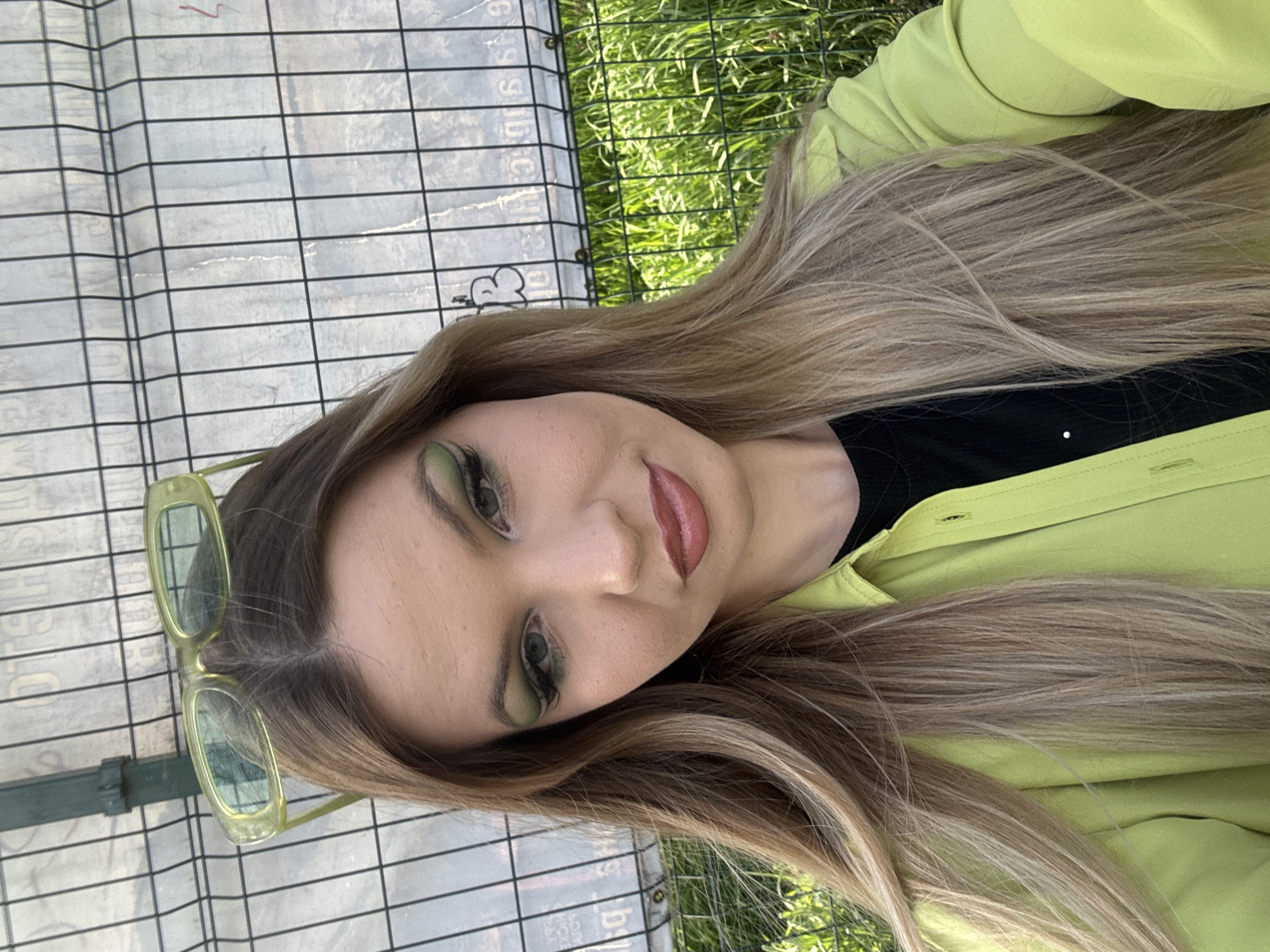 A young woman is pictured from a high angle in Plovdiv, Bulgaria, wearing a neon green button-down shirt over a black top. She has long, blonde hair with dark roots and green eyeshadow. Neon green sunglasses sit atop her head. Behind her is a fenced area containing a wire grid and green grass.
