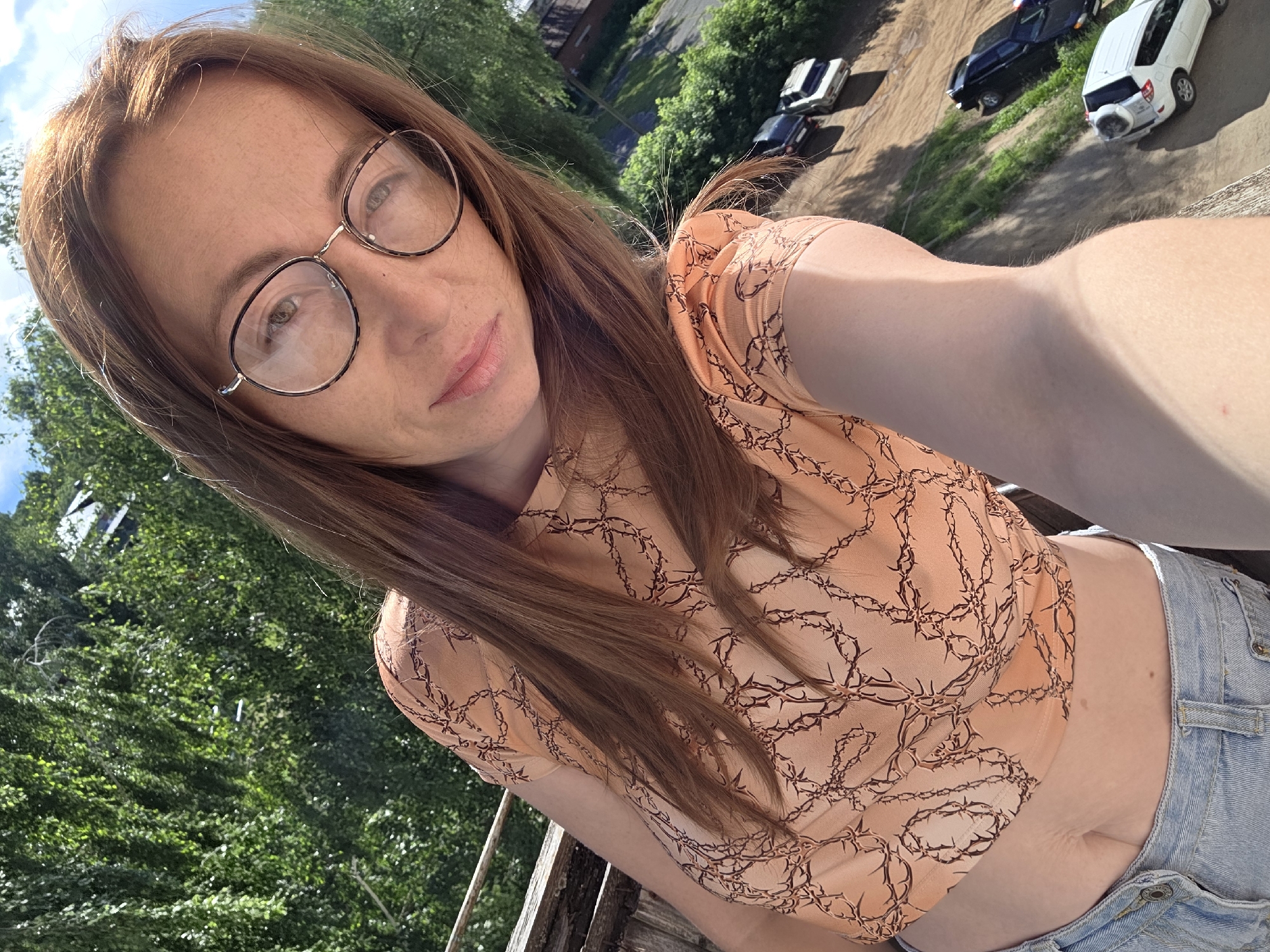 In this selfie, a woman with long, auburn hair and glasses is pictured outdoors. She is wearing a light peach-colored crop top with a brown, barbed wire-like pattern and light blue denim shorts. The woman appears to be looking towards the camera with a slight smile.
The background reveals a scene with trees and several cars parked on a dirt road. The sunlight suggests it is daytime, and the foliage indicates a warm season. The perspective of the photo, taken from an elevated position, hints that the woman might be on a balcony or a higher level overlooking the street. The overall impression is a casual, candid moment captured in an outdoor setting.
