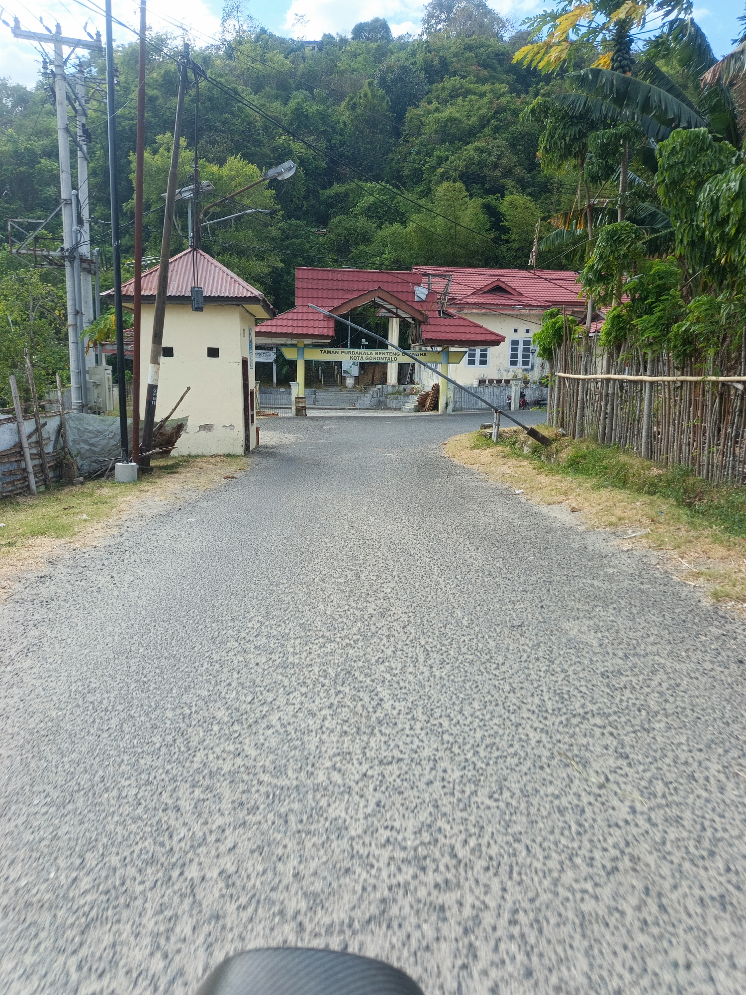 The image shows a paved road leading towards a building complex with red roofs, possibly an archaeological site or historical complex, as indicated by a sign that reads "TAMAN PURBAKALA BENTENG O NAPA KOTA GORONTALO". The complex is surrounded by lush green trees and vegetation, with a hillside covered in dense forest in the background. Overhead power lines are visible on the left side of the road, supported by concrete poles. A small cream-colored building with a red roof stands to the left of the entrance to the complex. The road surface is asphalt with visible gravel. The sky is partly cloudy.
