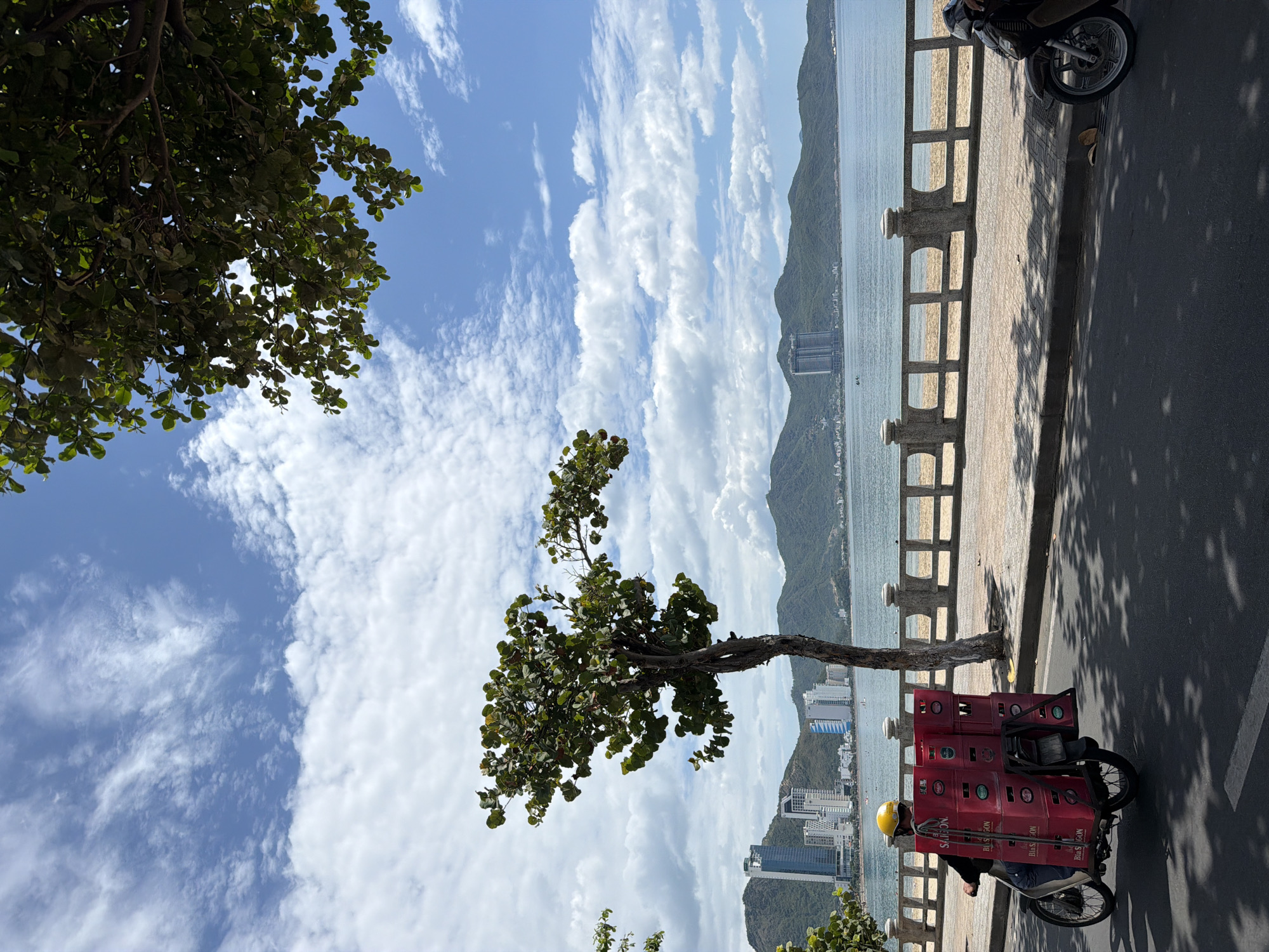 A man wearing a yellow helmet rides a motorcycle on a street beside a beach in Nha Trang, Vietnam. The motorcycle has a rack on the back carrying stacked red crates with the "BIA SAIGON" logo. The street has a paved sidewalk with a decorative stone railing overlooking a sandy beach. Beyond the beach is the calm sea and a coastline with lush green mountains in the background, dotted with tall buildings and a few smaller structures. The sky is bright blue with scattered white clouds. The scene appears to be during daylight, with shadows cast on the street from trees lining the sidewalk.