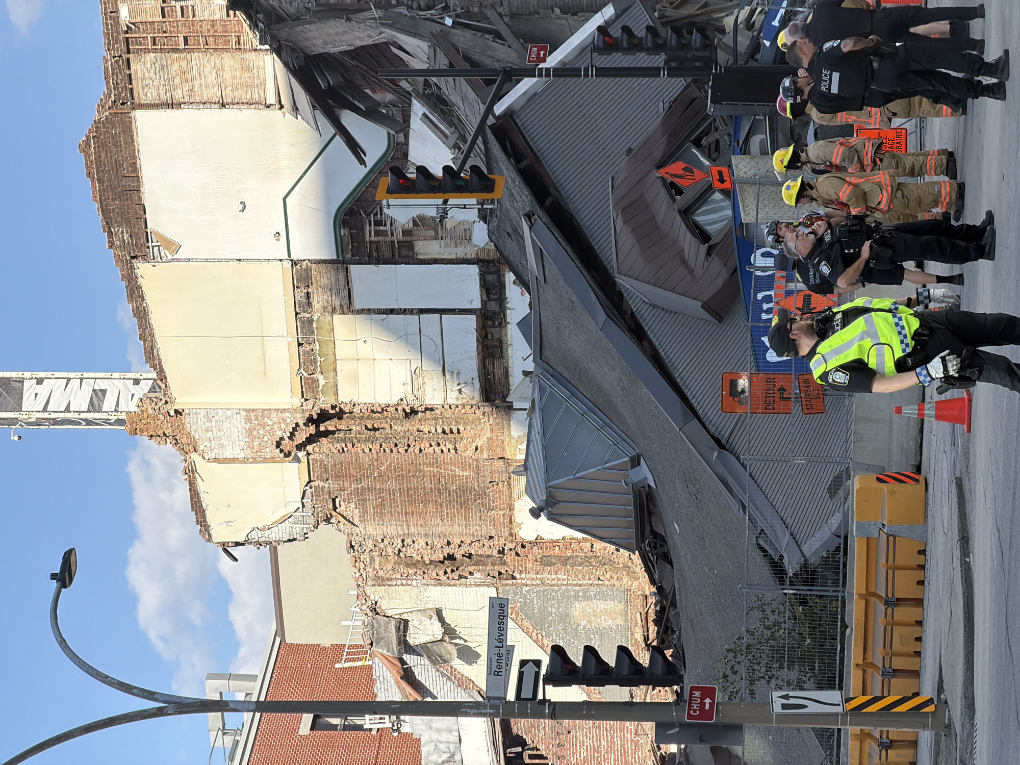 The image captures a scene of structural damage and emergency response in Montreal, Canada. In the background, a partially collapsed building reveals exposed brickwork and debris, with a construction crane visible above. The foreground is occupied by a group of individuals in uniform, including police officers and firefighters, indicating an ongoing emergency operation. Traffic lights and street signs, one reading "René-Lévesque," are also present, suggesting the location is an intersection. The overall environment appears to be an urban setting, with elements of construction, demolition, and public safety personnel engaged in their duties. The scene is orderly, with personnel appearing to be coordinating and assessing the situation.