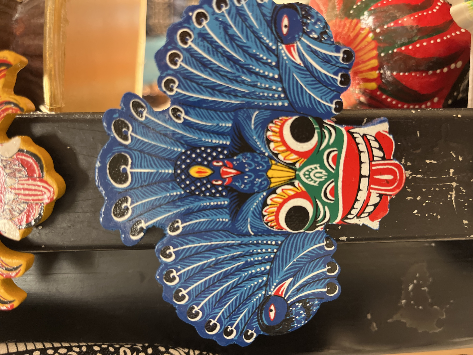 The image displays a close-up, eye-level shot of a vibrant, intricately painted mask, likely a decorative piece, possibly found in Katunayake, Sri Lanka. The mask is predominantly blue, green, red, and yellow, with black accents. It features a central, wide, green-faced figure with large, red-rimmed eyes and a gaping red mouth revealing white teeth and a red tongue sticking out. Flanking the central face are stylized peacock feathers in various shades of blue, detailed with white lines and black circular markings. Two smaller, blue bird-like figures with red beaks and red-rimmed eyes are positioned on either side, within the spread of the peacock feathers. The mask appears to be attached to a dark, upright surface, possibly a wooden post or frame, which shows signs of wear and tear with white specks and scratches. To the right of the mask, a portion of another colorful, round object, possibly a gourd or another decorative item, is visible. The background is blurred, but hints of a yellowish-orange hue suggest an indoor setting or a brightly lit area.