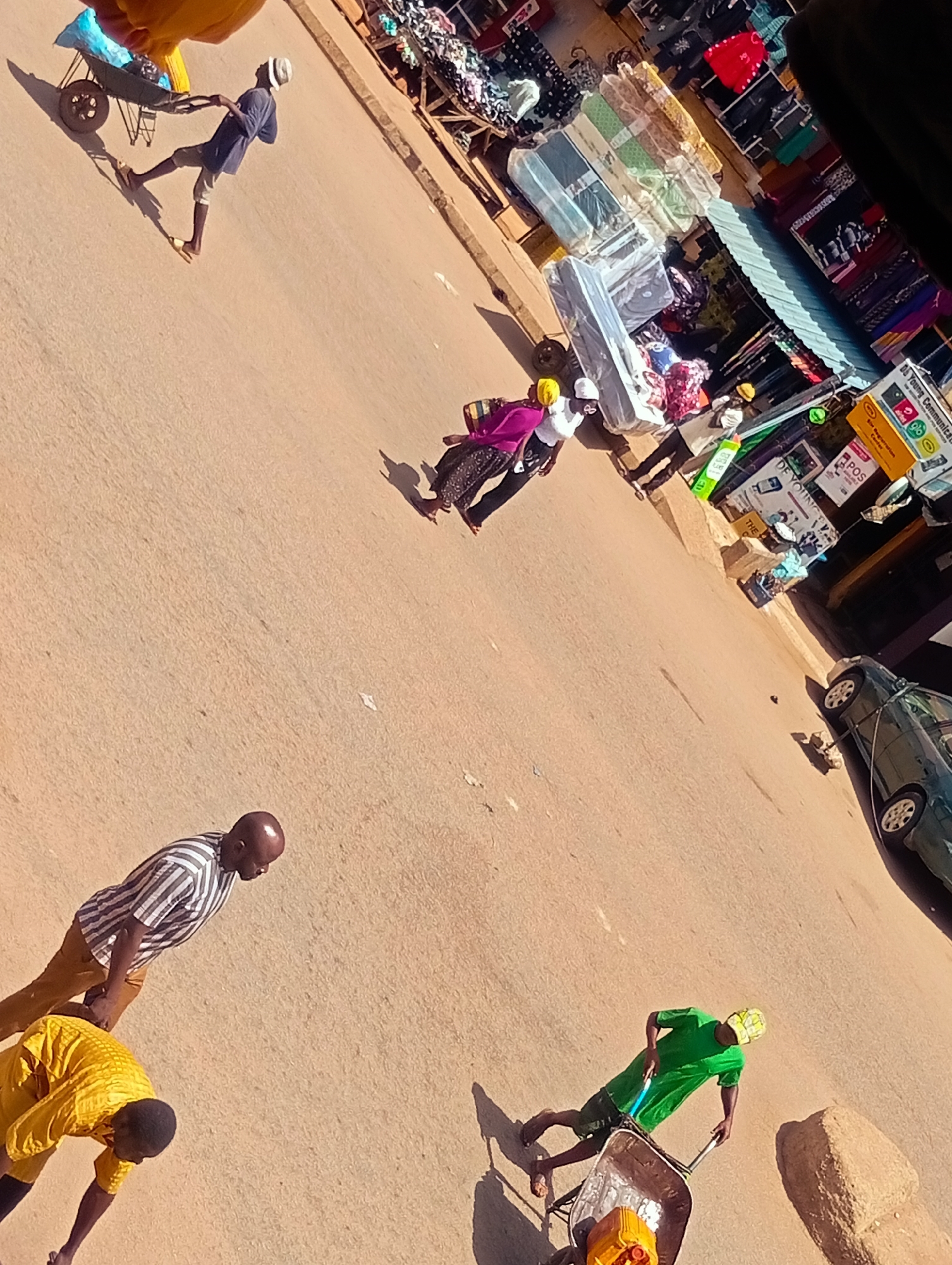 The image depicts a street scene in Bukuru, Nigeria. A person wearing a blue shirt and white hat is pulling a wheelbarrow loaded with blue and yellow items. Further down the street, a group of people are gathered around stalls displaying various goods, including mattresses and clothing. A dark green car is parked on the right side of the street. In the foreground, two men are walking on the street; one is wearing a striped shirt and brown pants, while the other is wearing a yellow shirt. Another person in a green shirt and checkered hat is pushing a wheelbarrow containing a yellow object. The ground is a dusty, reddish-brown color, indicative of a dry, possibly semi-arid environment. Sunlight casts distinct shadows, suggesting it is daytime.