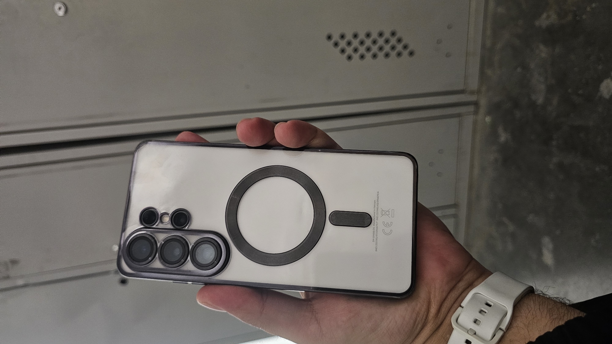 A person's hand, with visible wrist hair, is holding a white smartphone with a clear protective case. The back of the phone displays a circular magnetic charging ring with a smaller rectangular magnet below it. The camera module, featuring multiple lenses arranged in a vertical stack, is visible on the left side of the phone. The phone's casing has some subtle text and markings, including "SM-59488/DS Manufactured in Vietnam," "IMEI" numbers, and a "CE" logo. The background appears to be a metallic, textured surface, possibly lockers or shelving, with a row of small, dark circular perforations. The lighting suggests an indoor environment. No specific time of day or weather conditions can be determined.