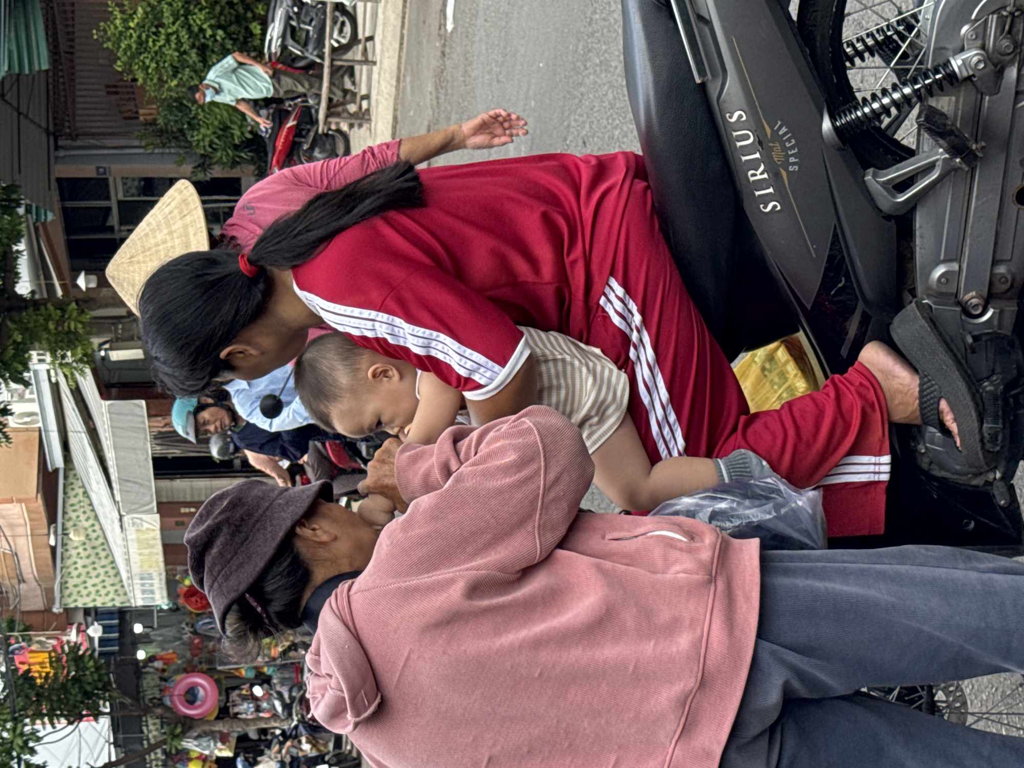 In Nha Trang, Vietnam, on a street, a woman in a pink hooded jacket and dark pants is handing a baby to another woman wearing a red tracksuit and a conical hat. The baby, wearing a striped shirt and pants, is held by the woman in red. A motorcycle is visible, with the word "SIRIUS" on its side. Other people and buildings can be seen in the background.