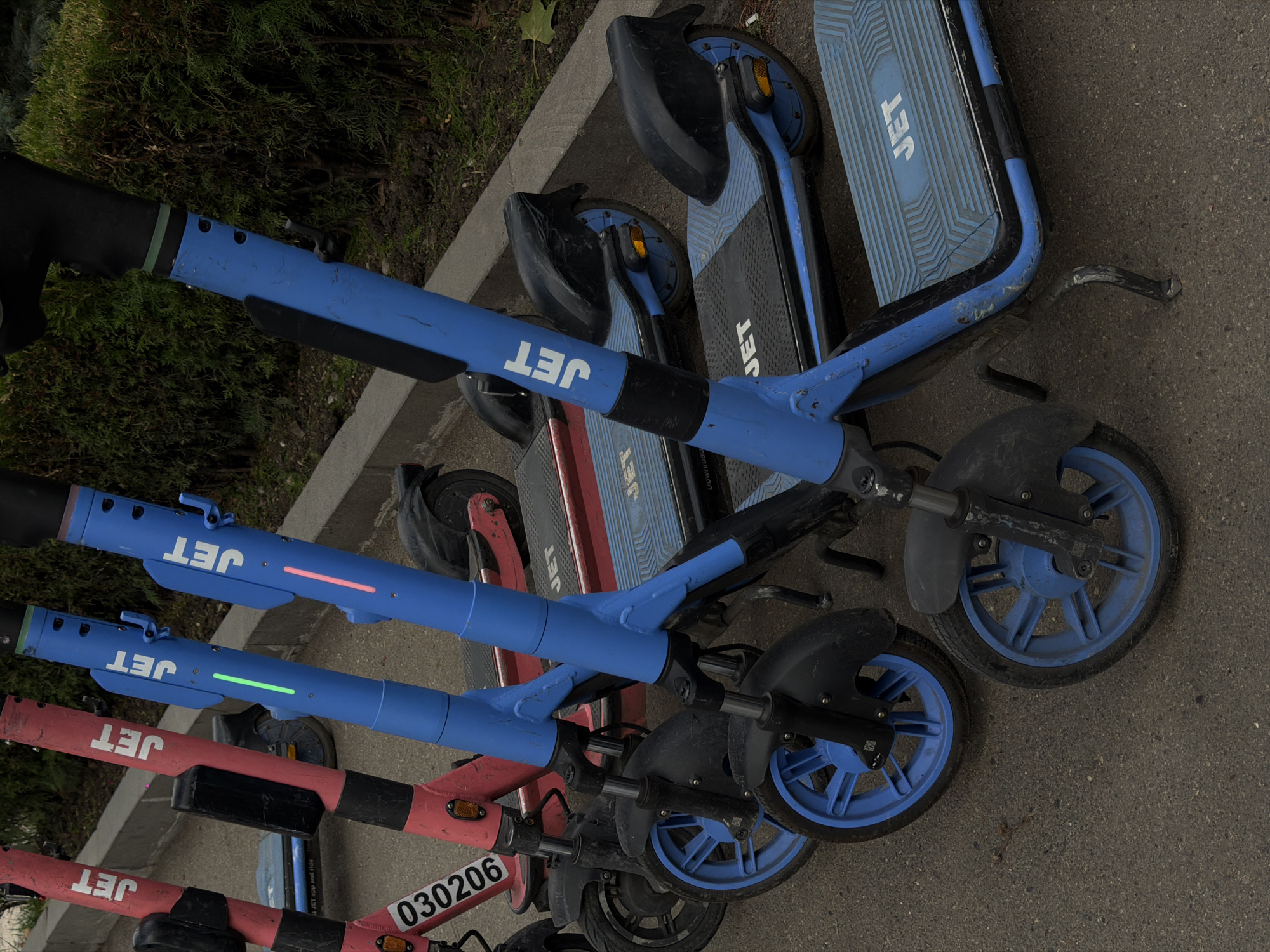 A row of parked electric scooters is visible, lined up on an asphalt surface next to a curb. The scooters are predominantly blue and red, with the "JET" logo prominently displayed on their decks and handlebars. Some of the blue scooters have illuminated vertical LED strips on their steering columns, one green and one pink. The scooters appear to be part of a rental service, as suggested by the number "030206" visible on the red scooter. In the background, dense green foliage forms a backdrop, indicating an outdoor setting. The overall scene suggests a common urban streetscape, likely a public area in Tbilisi, Georgia, where such shared mobility services are popular.