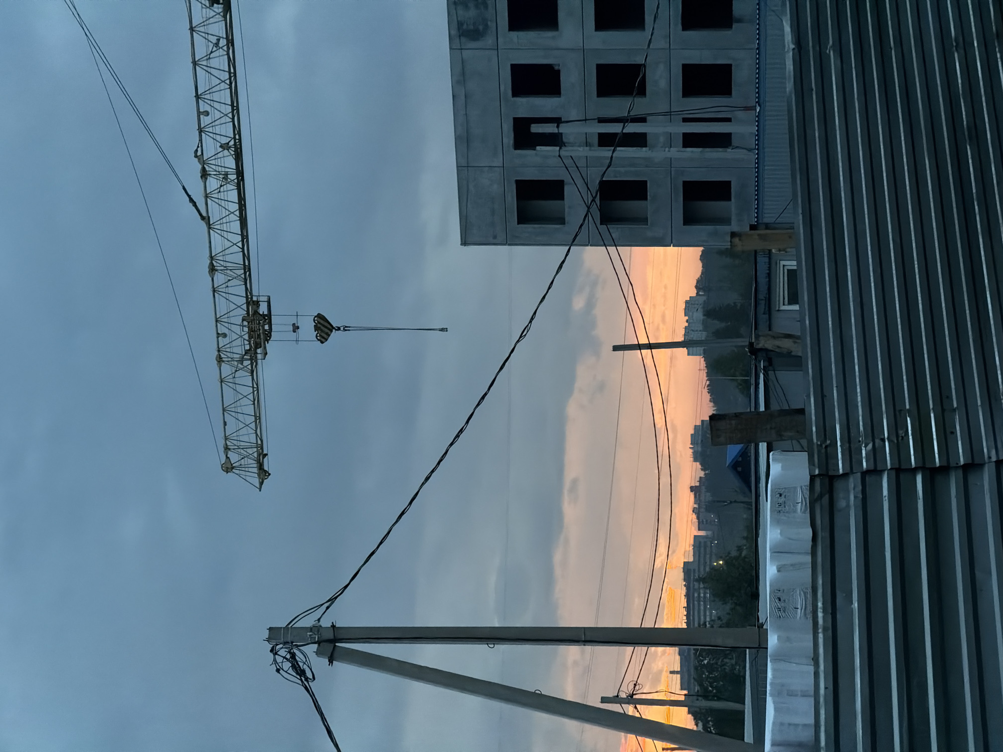 The image depicts a construction site at dusk in Froli, Russia. A tall yellow construction crane dominates the left side of the frame, its lattice structure silhouetted against a moody, overcast sky. The crane's boom extends diagonally upwards, with a hook mechanism visible at its end. In the background, a partially constructed concrete building with rows of dark, empty window frames stands out against the sky. A corrugated metal fence, likely part of the construction site's perimeter, runs vertically along the right edge of the image.
The sky displays a gradient of colors, transitioning from a deep, cloudy grey at the top to a warm, soft orange and pink hue near the horizon, indicative of sunset. This warm glow illuminates the distant cityscape, where the outlines of apartment buildings and trees are faintly visible. Power lines crisscross the scene, supported by a concrete utility pole in the foreground, adding to the urban industrial atmosphere.
The overall mood is one of quiet anticipation and the end of a day's work. The dimming light and the presence of the unfinished building suggest a pause in activity, with the construction project awaiting completion. There is no visible text in the image.