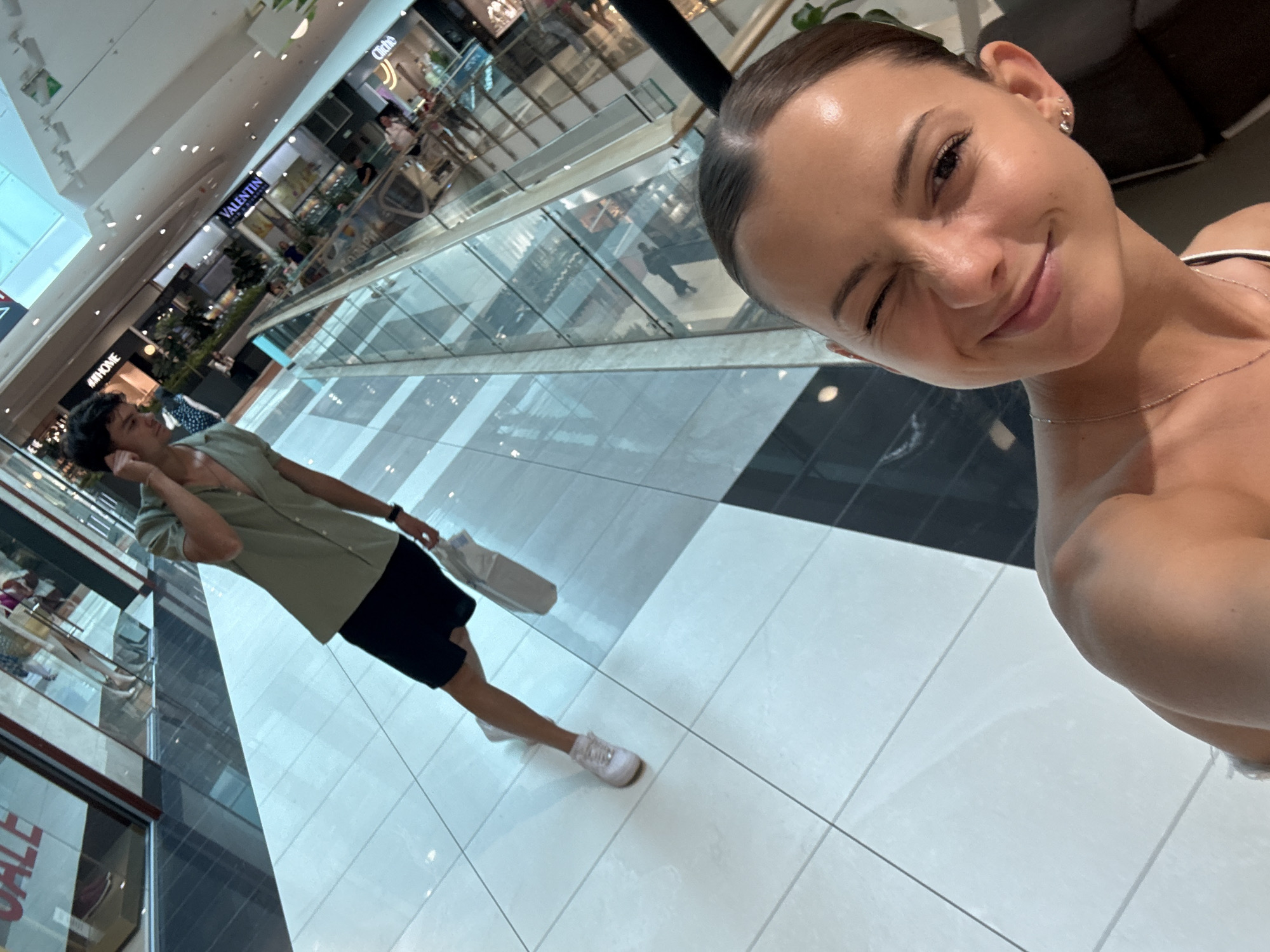 The image captures a young woman taking a selfie with a wink in the foreground, her right shoulder and upper chest visible. She is wearing a thin necklace and small earrings. In the background, a young man walks away from the camera, holding a white shopping bag and a phone to his ear. He is dressed in a light green short-sleeved shirt and black shorts, with white sneakers. The scene takes place in a modern shopping mall with a tiled floor and glass railings overlooking multiple levels. The mall appears to be in Sofia, Bulgaria, suggested by storefront signage that includes the name "VALENTIN" and other indistinct brand names. The lighting is bright, characteristic of an indoor retail environment.