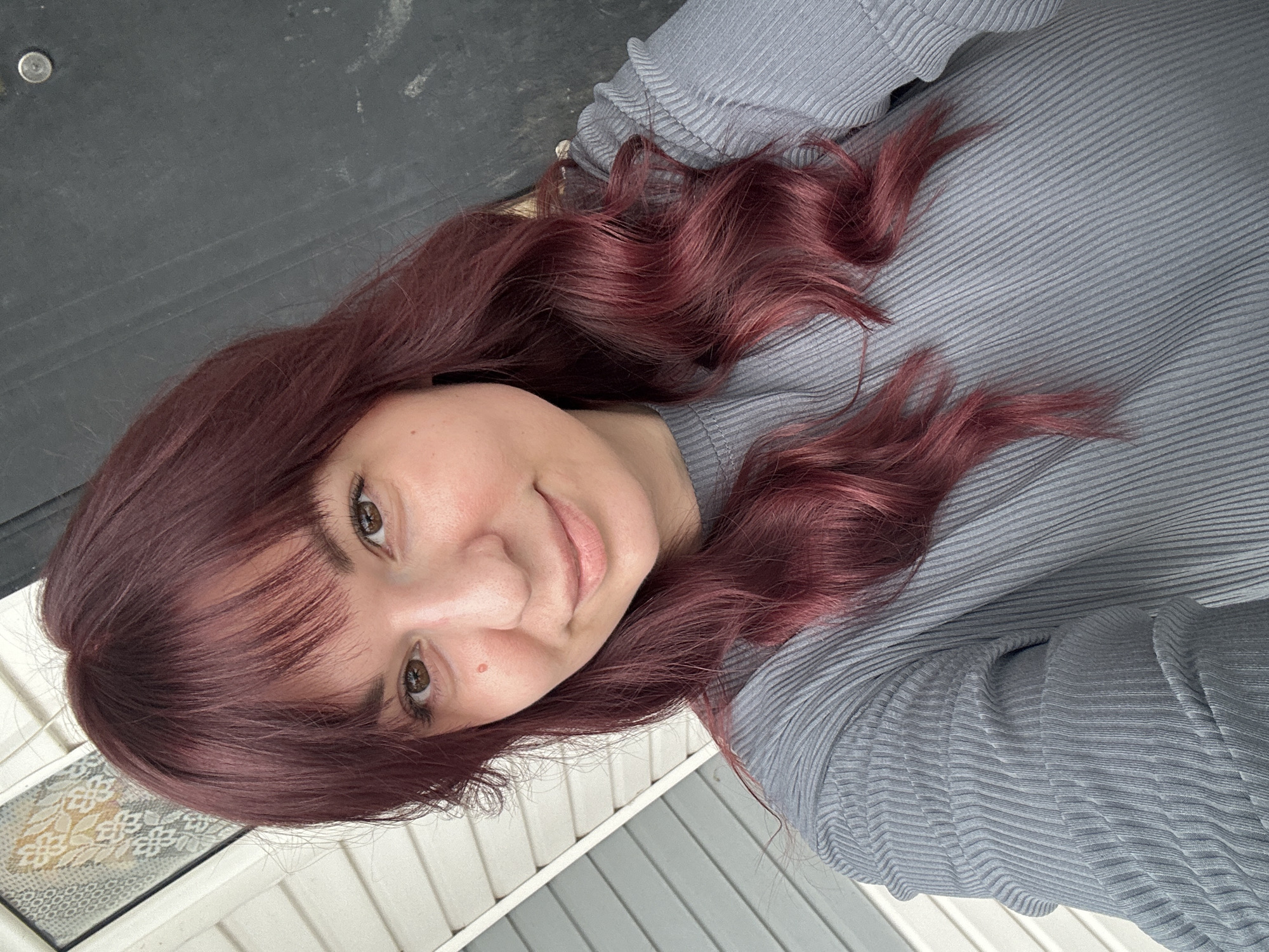 A young woman with shoulder-length, wavy, maroon-colored hair and bangs is depicted from a high angle, likely in a selfie. She is wearing a grey, ribbed long-sleeved shirt. The background includes a grey-painted surface, possibly a door or wall, and a white, horizontally paneled surface, possibly siding, with a doormat featuring a diamond pattern visible at the bottom left. The location Фоки, Russia, cannot be determined from the visual elements.