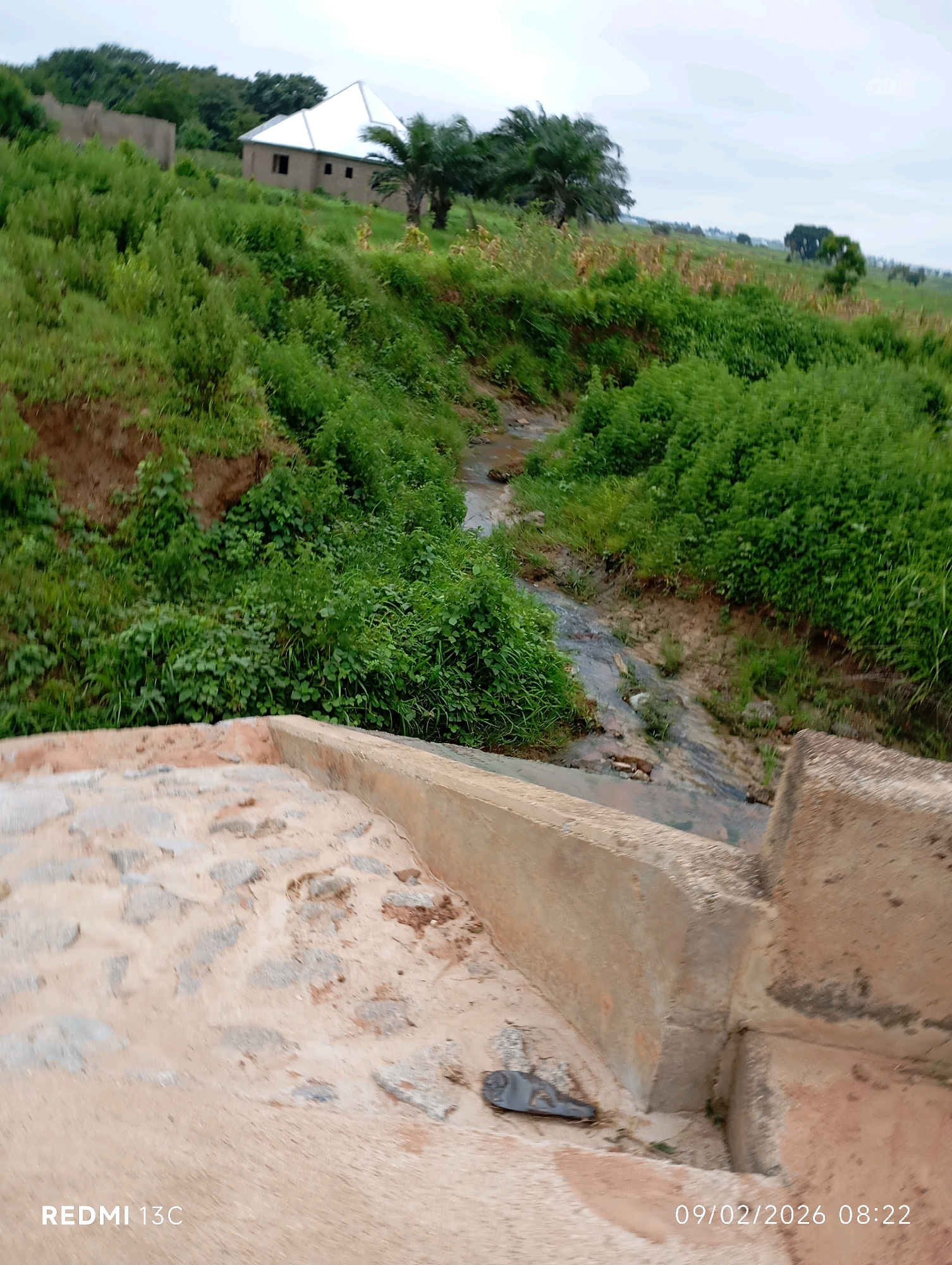 The image depicts a partially constructed building with a white metal roof situated on a lush green hillside in Shendam, Nigeria. A small, winding stream flows through the dense vegetation that covers the landscape. In the foreground, a concrete structure, possibly a bridge or embankment, is visible, with a rough, stone-like surface and worn concrete edges. A dark, irregularly shaped object, resembling a shoe or a piece of debris, rests on the concrete in the lower left of the frame. The sky is overcast. The date and time stamp "09/02/2026 08:22" and "REDMI 13C" are overlaid on the image.