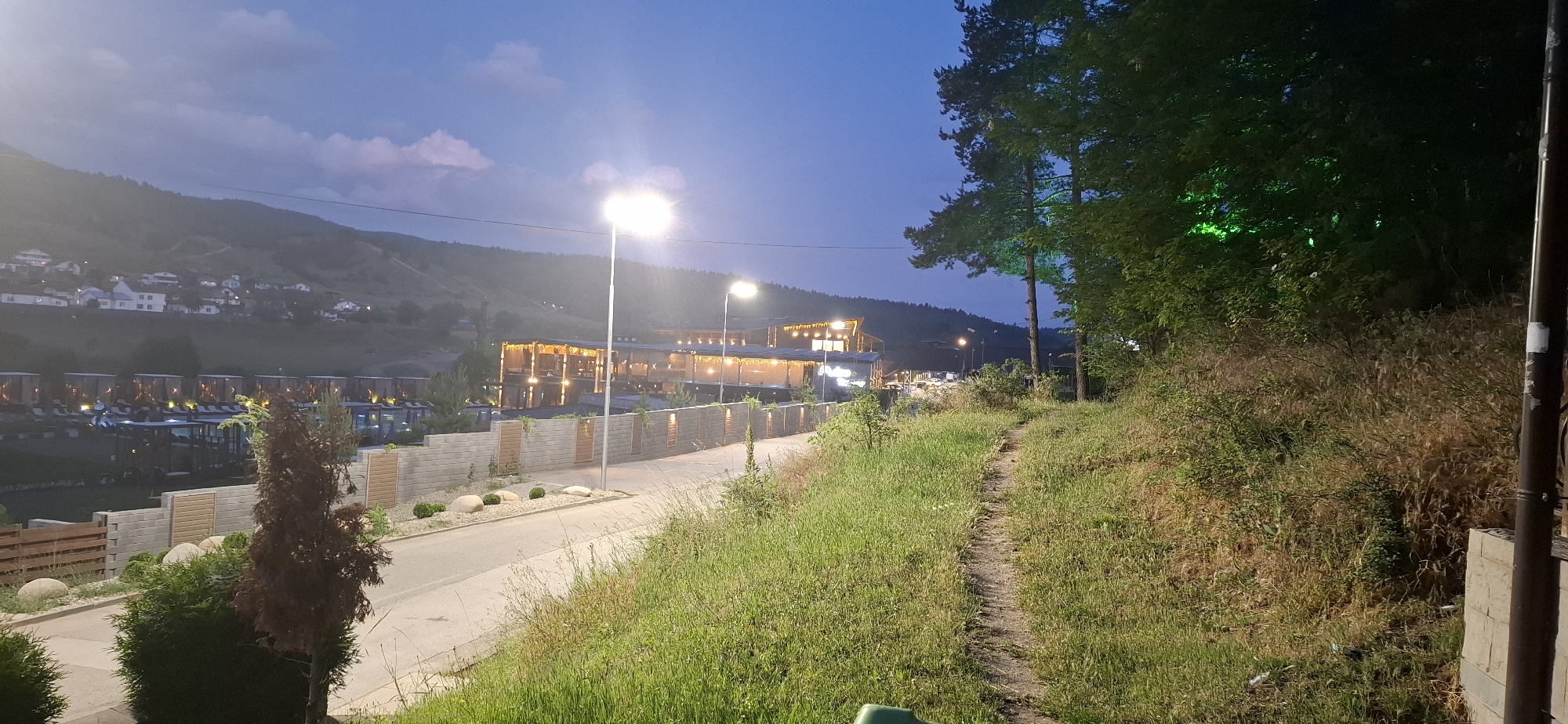 This detailed description pertains to an evening scene in Bania, Bulgaria, captured at twilight.
The image presents an outdoor landscape at dusk, characterized by a deep blue sky with faint cloud formations. In the foreground, on the right, a narrow dirt path ascends a grassy, uneven slope covered with a mix of green and dry vegetation. Several trees with dark foliage stand on the right, two of which are noticeably illuminated with green accent lighting. A dark, vertical pole is partially visible on the far right.
To the left of the dirt path, a paved asphalt road curves gently to the left and recedes into the distance. Multiple streetlights with bright white illumination are positioned along the road. A constructed barrier, consisting of horizontal rows of grey concrete blocks interspersed with vertical wooden panels, runs parallel to the road on its left side. Landscaped areas in front of this wall feature rounded stones and low-lying green bushes. Closer to the viewer on the left, a small, conical evergreen tree with reddish-brown foliage and a cluster of green bushes are present.
Beyond the road and wall, a complex of modern buildings is visible. These structures are extensively illuminated with warm, orange-toned lights, likely from interior lighting and decorative string lights. A large area within the complex displays numerous outdoor seating arrangements, including lounge chairs, and several pool-like structures, some covered by dark canopies. Bright white lights also punctuate this area.
In the mid-to-far background, rolling hills rise prominently against the twilight sky. Numerous small, predominantly white houses with dark roofs are scattered across these hillsides, some exhibiting interior illumination. No individuals are discernible within the frame.