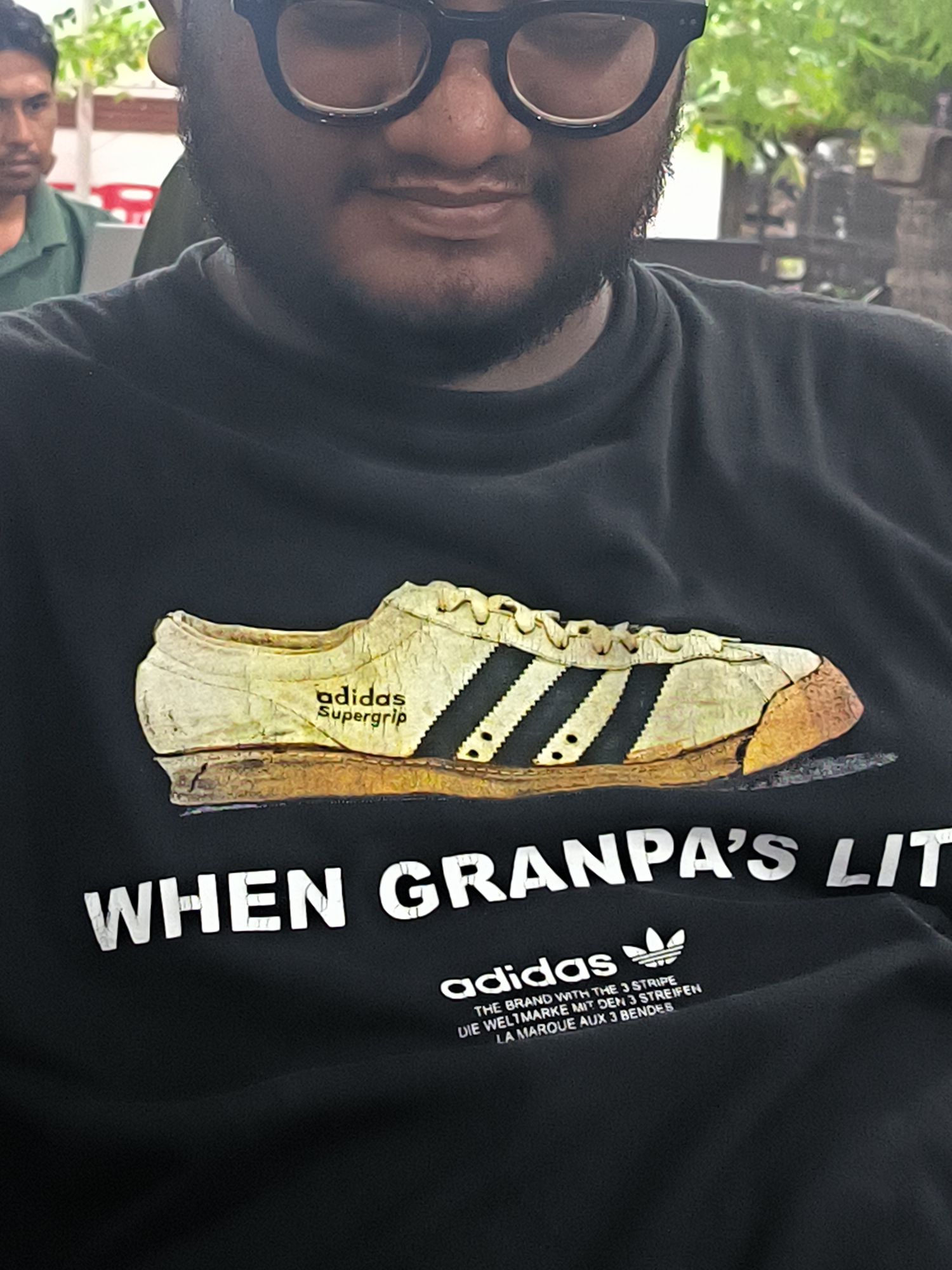 A close-up, chest-up shot features a man with a dark beard and black-framed glasses, captured with a subtle smile. He is prominently wearing a black t-shirt adorned with a graphic of a vintage, worn-out Adidas Supergrip sneaker. The shoe displays signs of age, with cracked textures on its light-colored upper and a scuffed, brownish sole, complemented by the classic three black stripes.
Below the sneaker graphic, large white text on the t-shirt reads: "WHEN GRANPA'S LIT". Further down, the Adidas logo is present, followed by multilingual branding slogans: "THE BRAND WITH THE 3 STRIPE", "DIE WELTMARKE MIT DEN 3 STREIFEN", and "LA MARQUE AUX 3 BENDES".
In the soft-focus background, another man in a green polo shirt is partially visible to the left. Blurred elements like green foliage and a red chair suggest an outdoor or semi-outdoor casual setting, possibly a cafe or eatery, in Lhokseumawe, Indonesia. The lighting indicates it is daytime. The man's relaxed posture and slight smile contribute to a casual and pleasant mood.